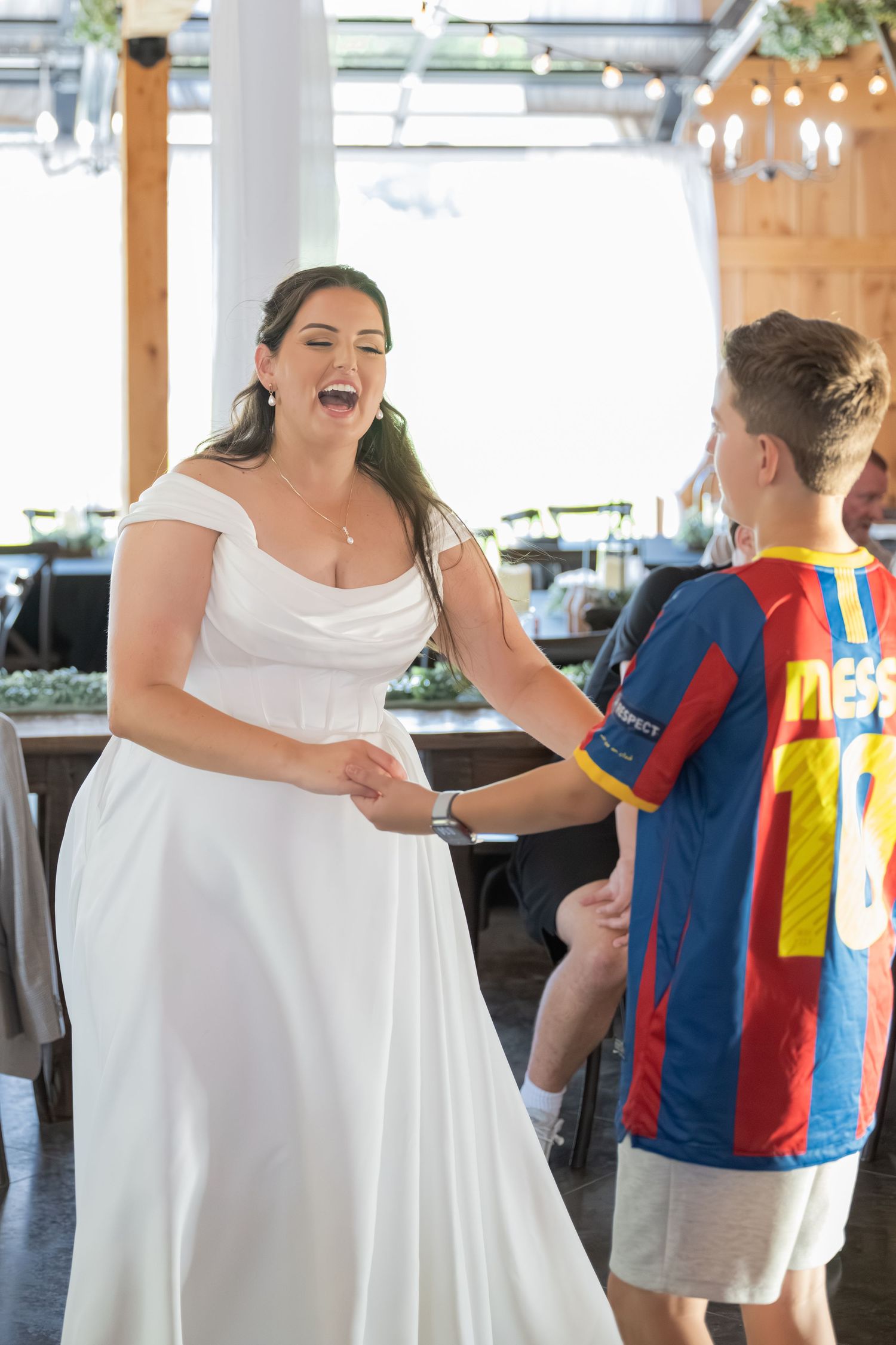 Bride in white gown laughing with young boy in Messi soccer jersey at wedding reception.