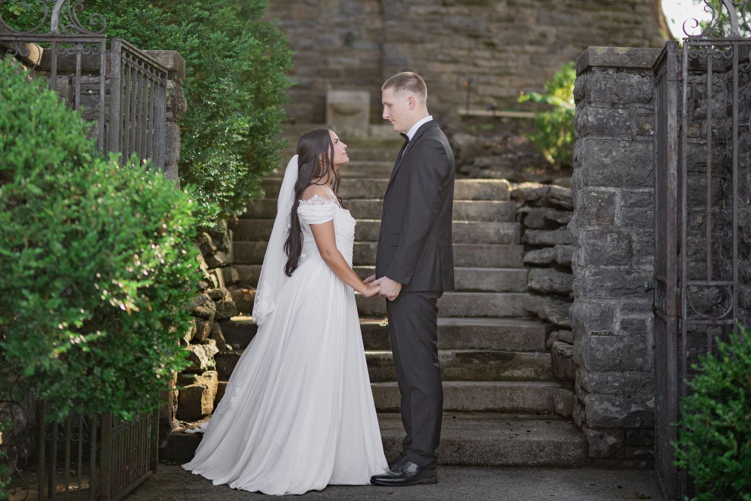 Bride and groom holding hands on stone steps outdoors, bride in white gown with veil, groom in dark suit.