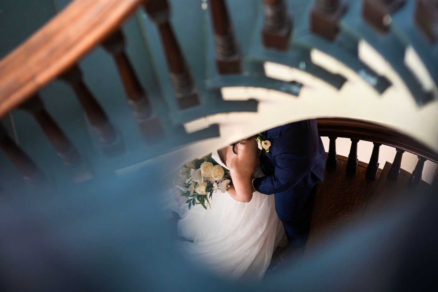 Aerial view of bride and groom embracing on a wooden spiral staircase, bride holding a white floral bouquet.