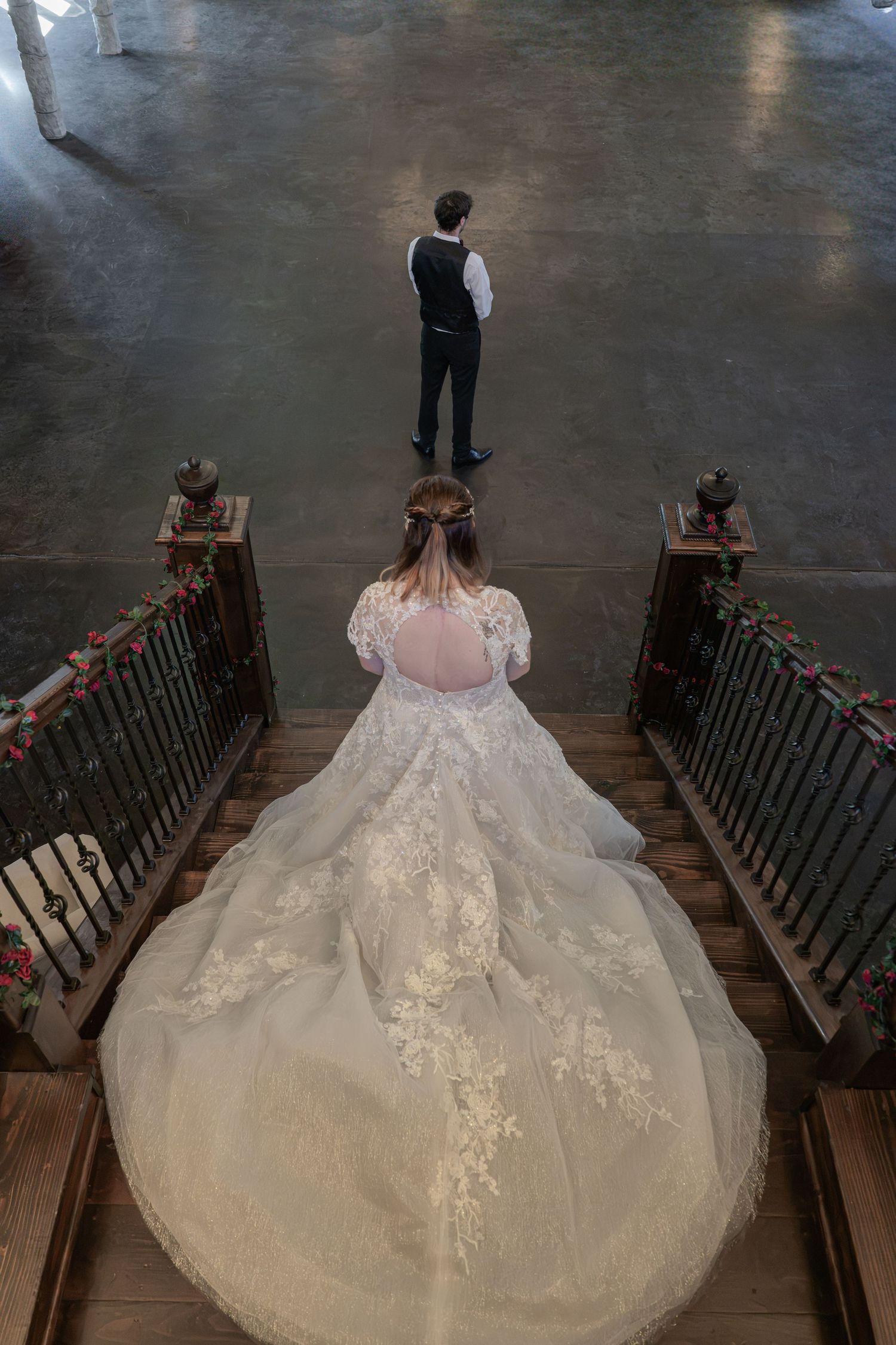 Aerial view of bride in lace gown descending staircase toward groom in black vest at wedding venue.
