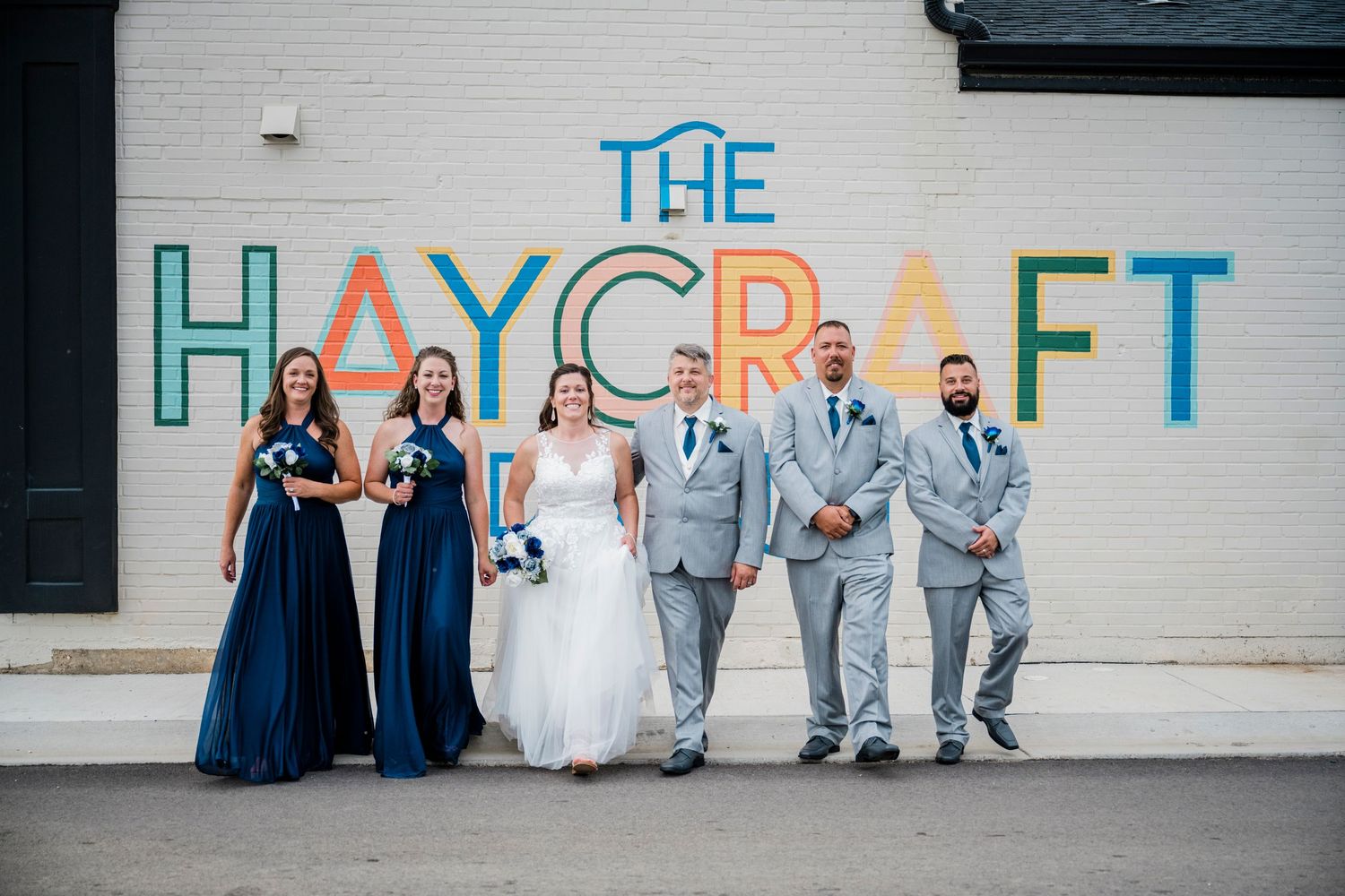 Wedding party poses before The Haycraft colorful mural, bridesmaids in navy gowns and groomsmen in light gray suits.