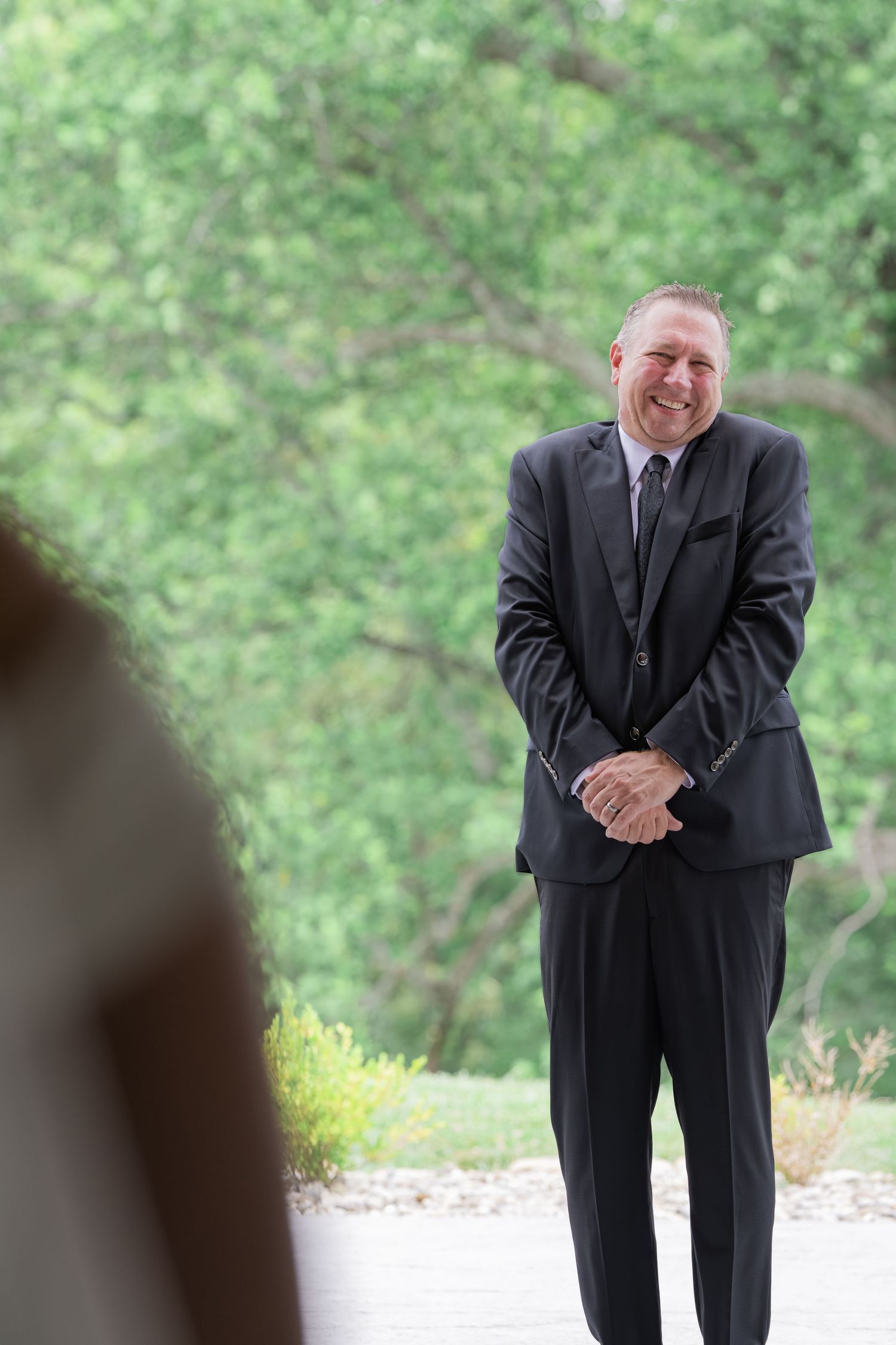 A smiling man in a dark suit with clasped hands stands outdoors at a wedding ceremony surrounded by lush green trees.