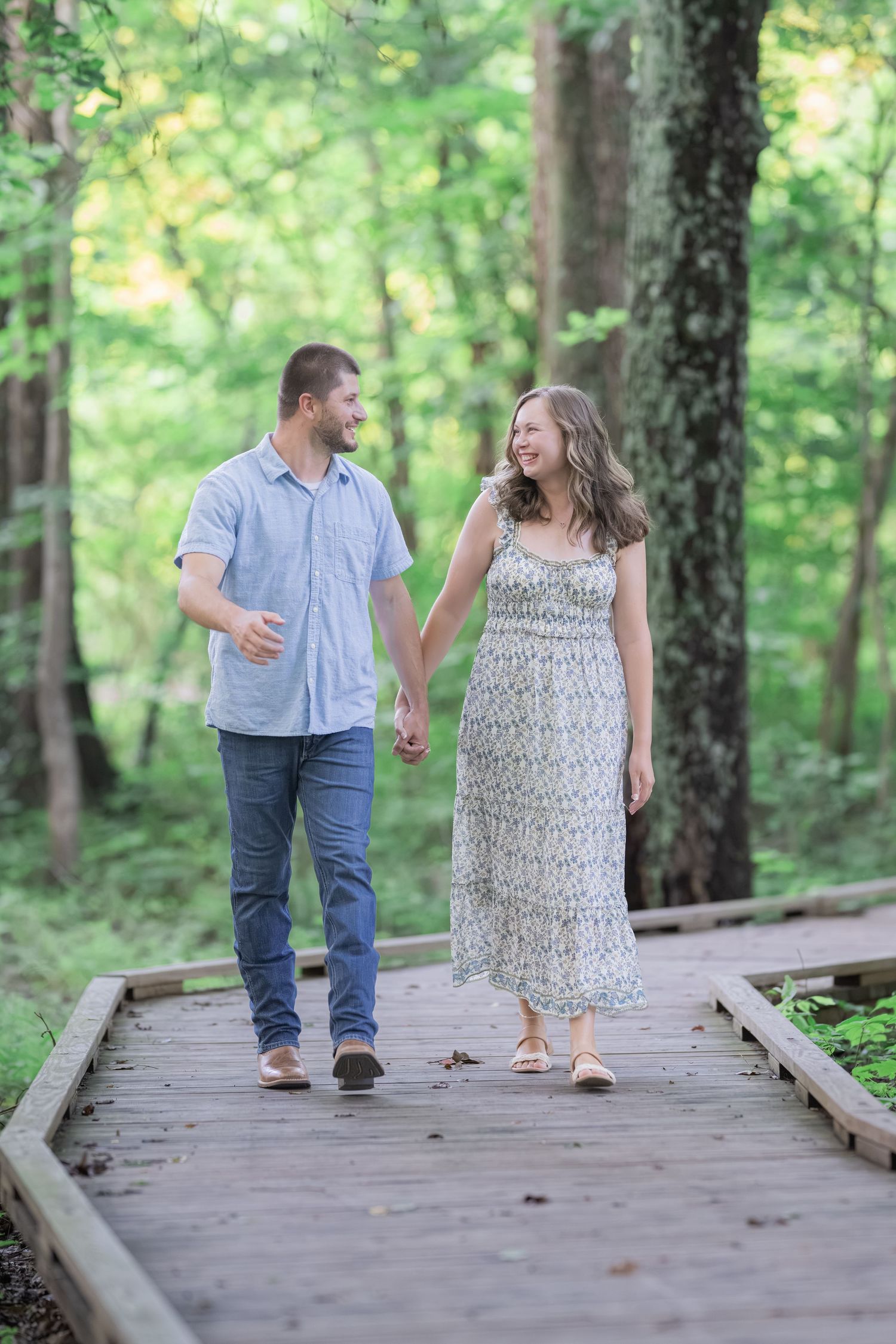 A couple holds hands and smiles while walking on a wooden boardwalk path through a lush green forest.