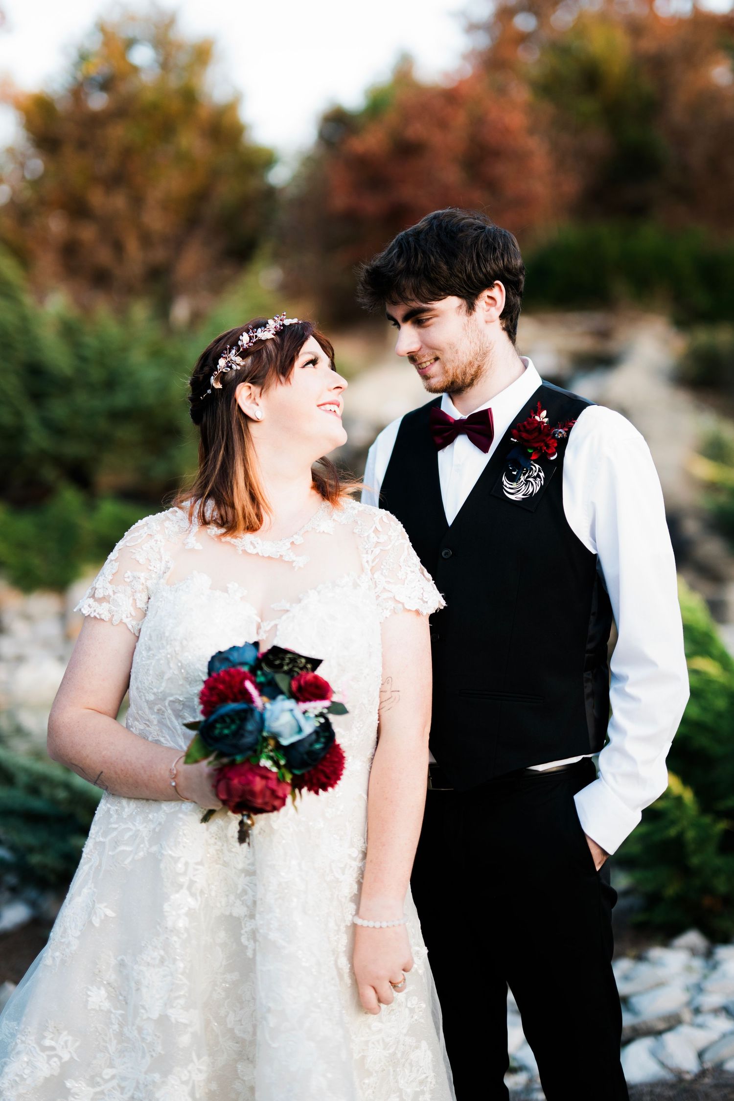 Bride in lace gown holding bouquet stands with groom in black vest and bow tie in an outdoor garden setting.