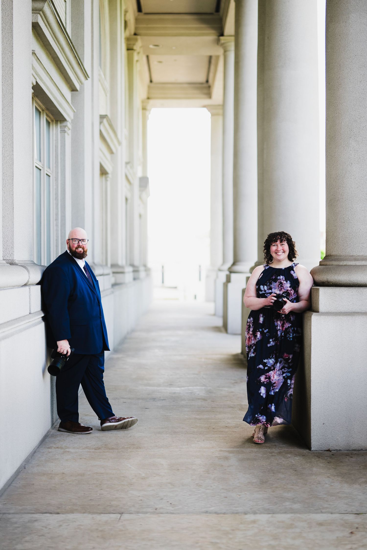 A man in a navy suit and woman in a floral dress pose between white columns of an elegant building.