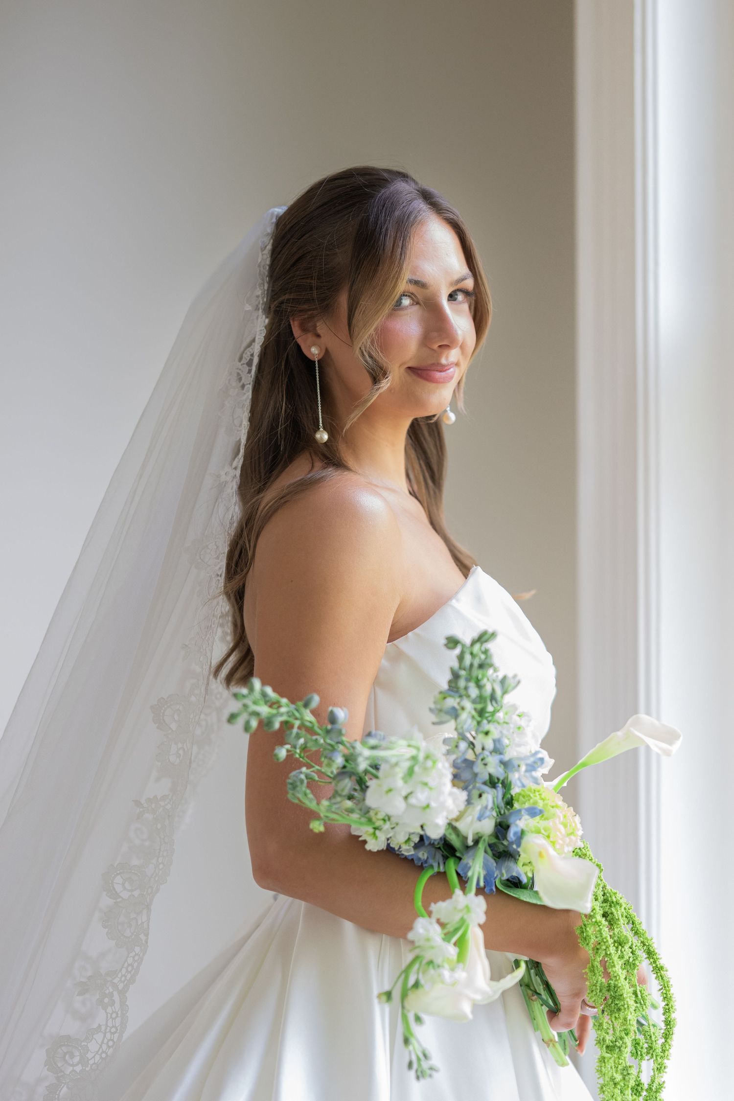 Elegant bride in white strapless gown and veil holds lush bouquet of white and blue flowers by a bright window.