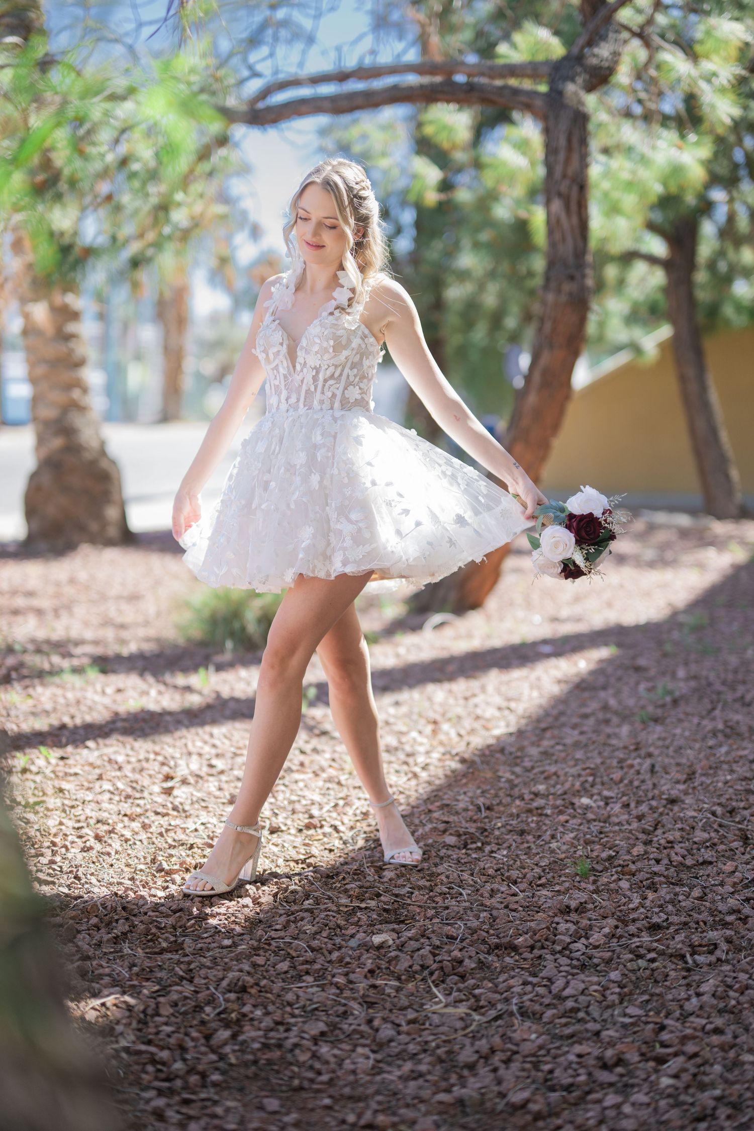 Bride in short white floral dress holding bouquet walks on gravel path surrounded by trees in outdoor setting.