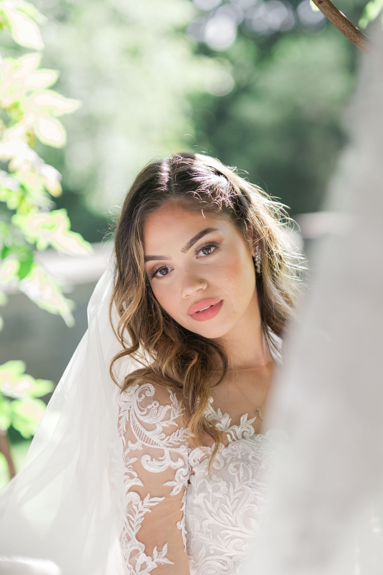 Bride in lace wedding gown and veil poses outdoors among green leaves in soft natural light.