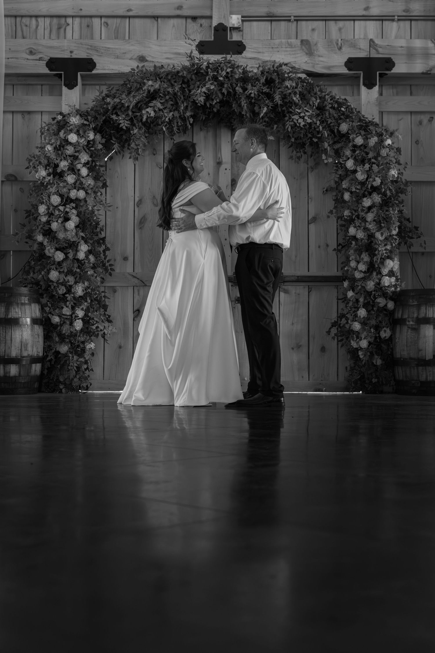 Black and white photo of bride and groom sharing first dance under floral arch in rustic barn venue.