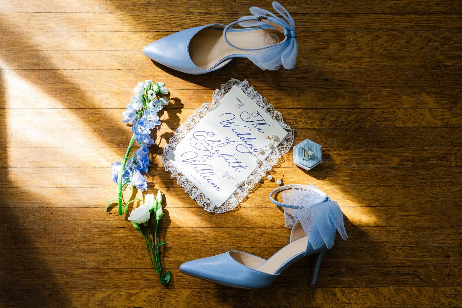 Blue bridal heels, floral bouquet, calligraphy vow book, and blue dice on wooden floor.