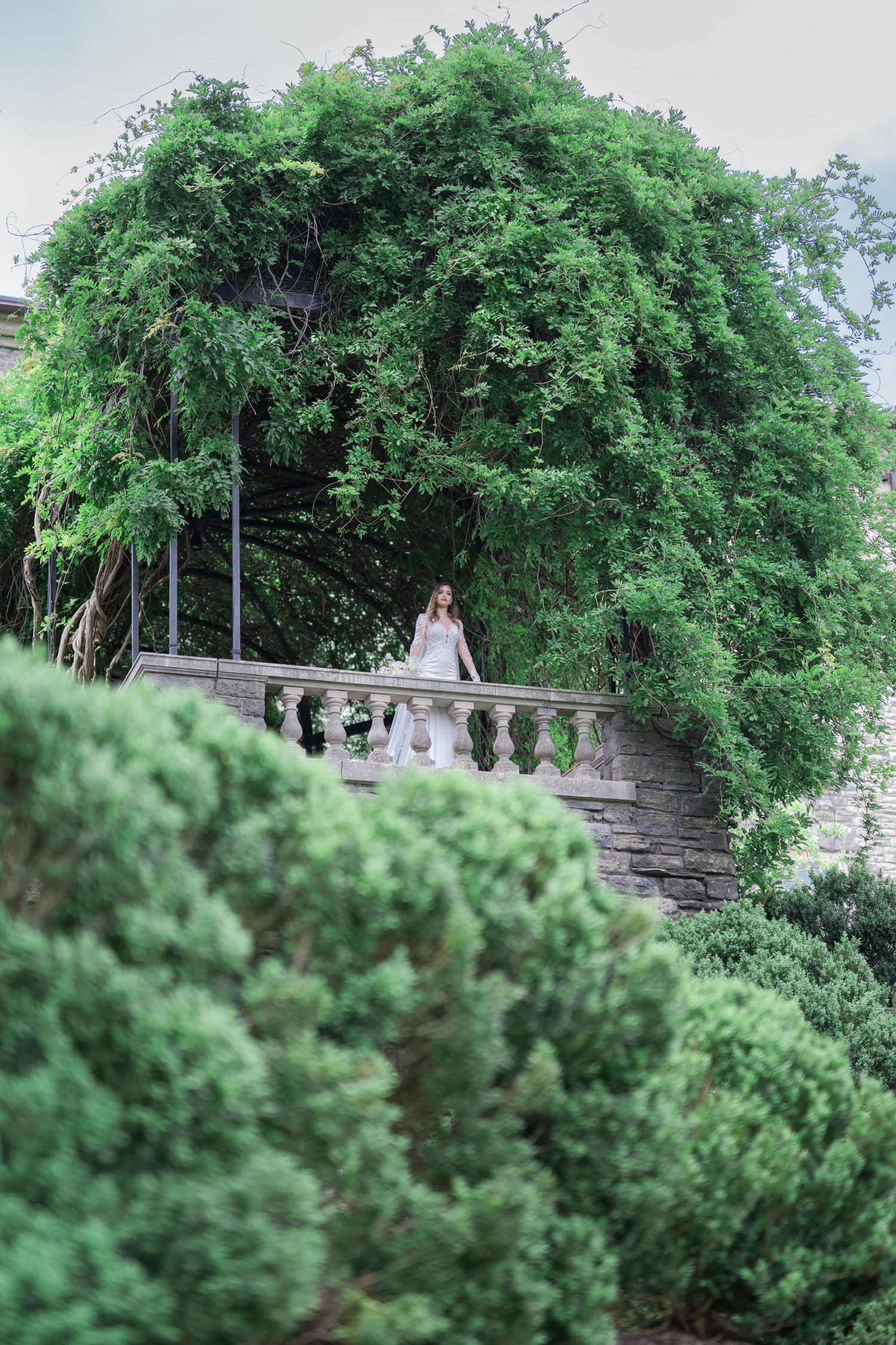 Bride in white gown standing on stone balcony surrounded by lush green trees and manicured shrubs in elegant garden.