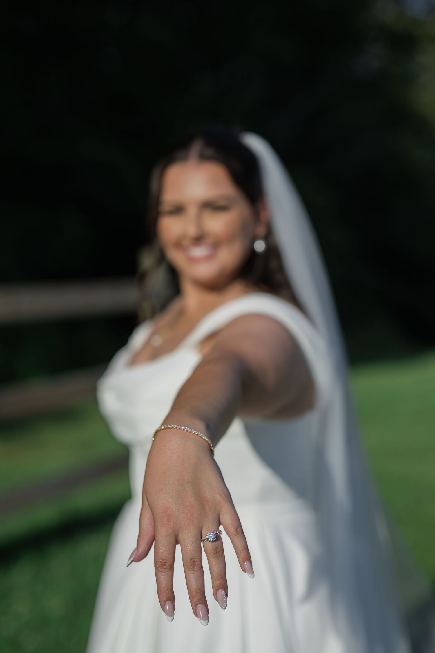 Smiling bride in white gown and veil extends hand toward camera showcasing elegant diamond engagement ring outdoors.
