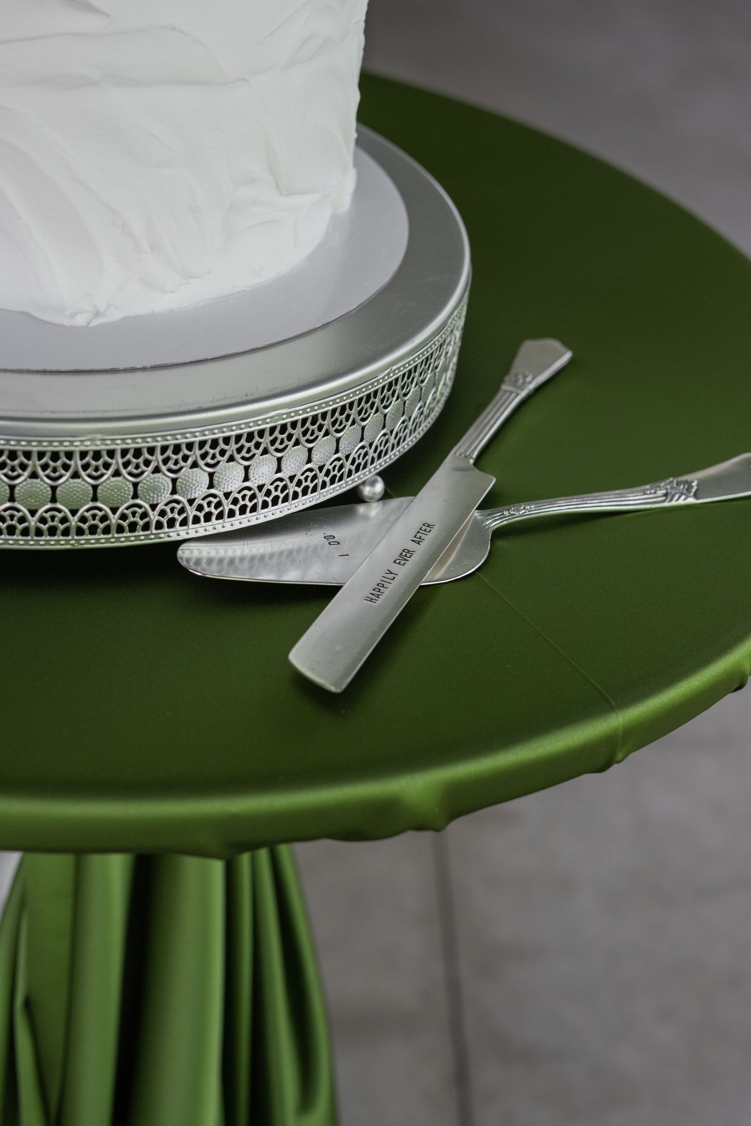 White wedding cake on silver stand with cake serving set on green draped table.