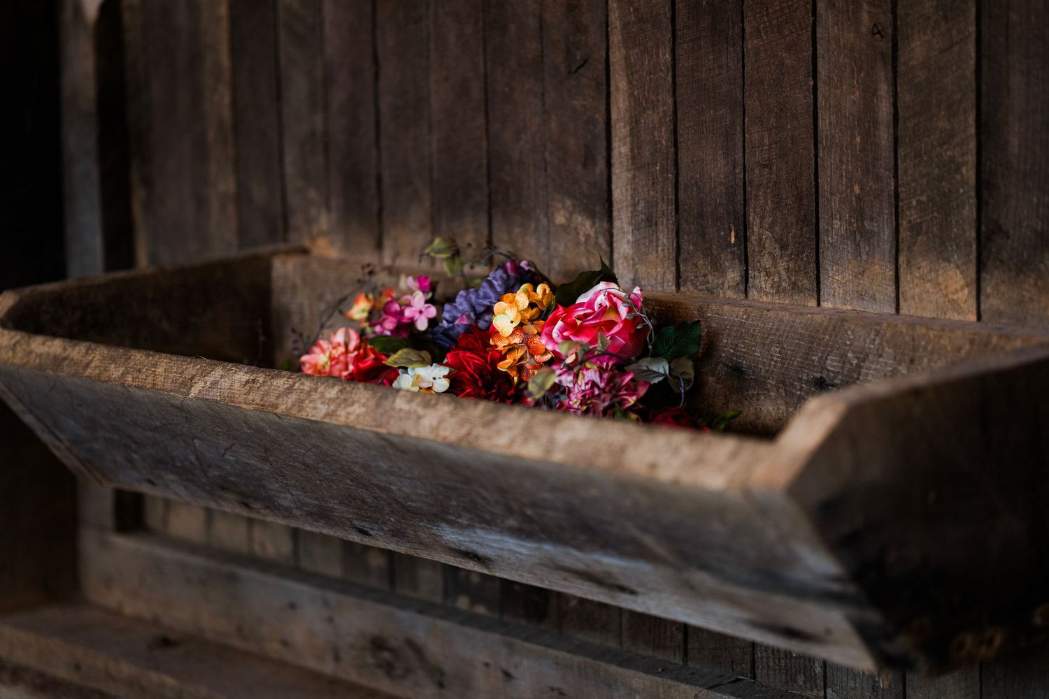 Colorful bouquet of pink, red, and orange flowers resting in a rustic wooden trough against a dark wood wall.