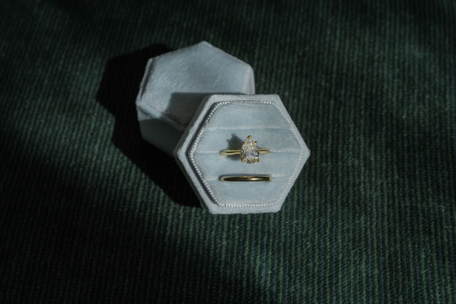 Gold diamond engagement ring displayed in a hexagonal white velvet ring box on a dark green textured surface.