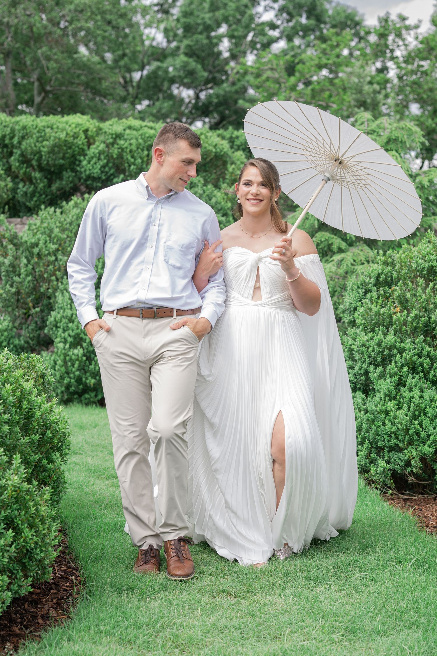 Bride in white gown holding parasol walks with groom in blue shirt through lush green garden path.