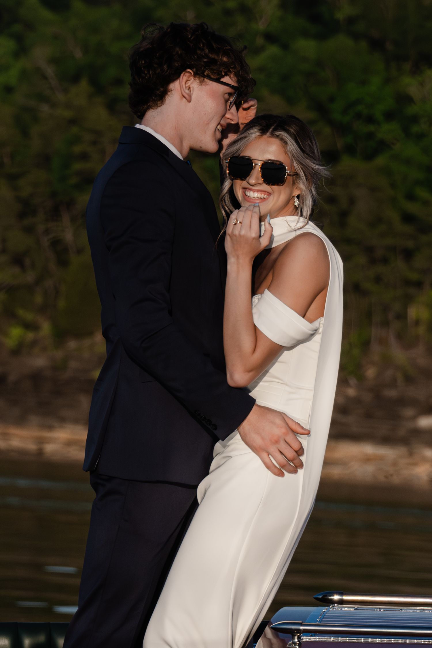 Couple in formal attire embracing on a boat, woman in white dress with sunglasses smiling, man in black suit.