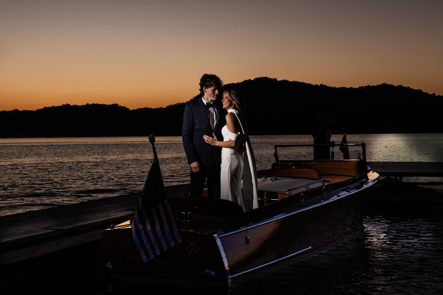 Couple embracing on a wooden boat at sunset with golden sky and silhouetted mountains over calm water.