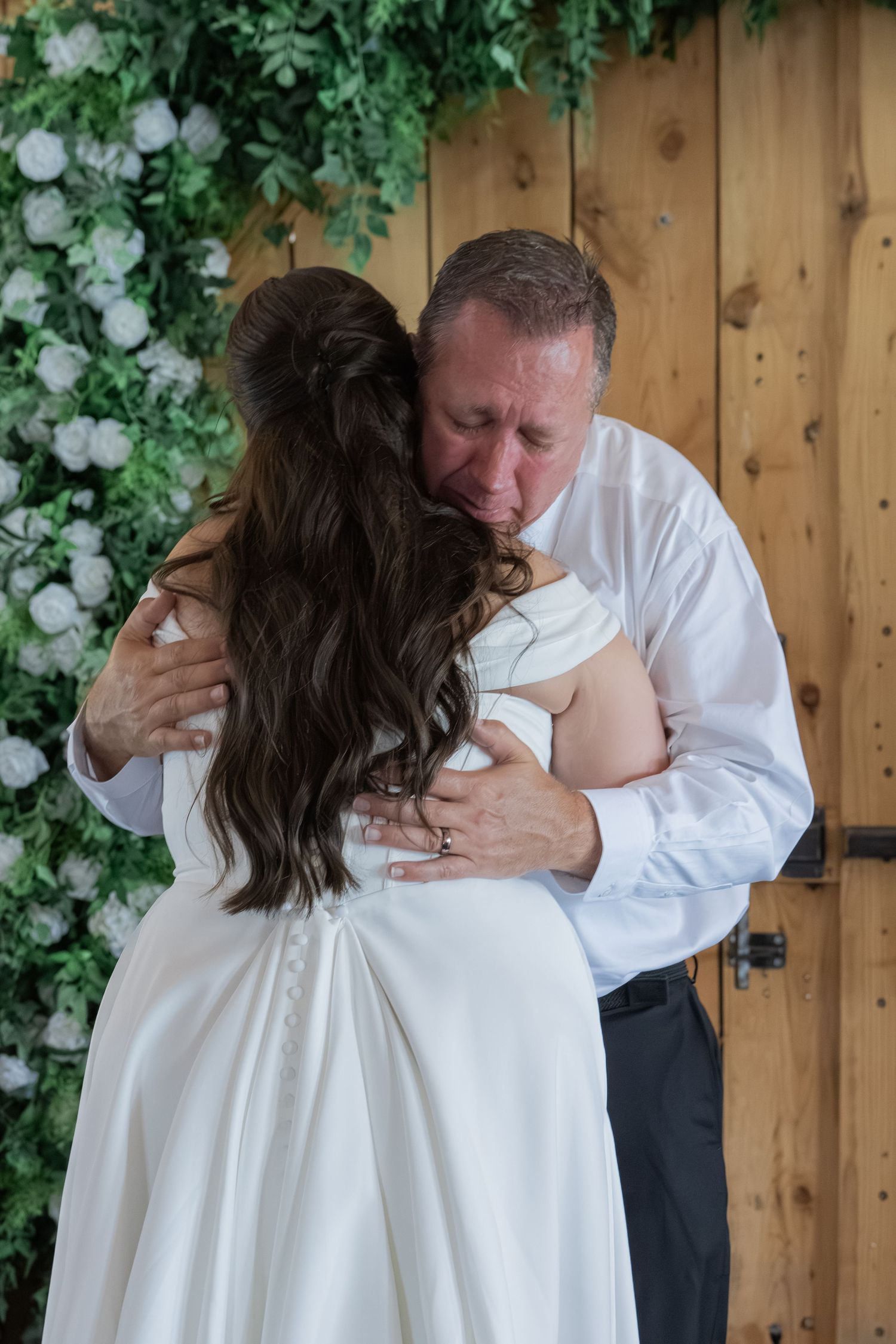 Father embracing bride in white wedding dress near floral wall and wooden barn doors on wedding day.