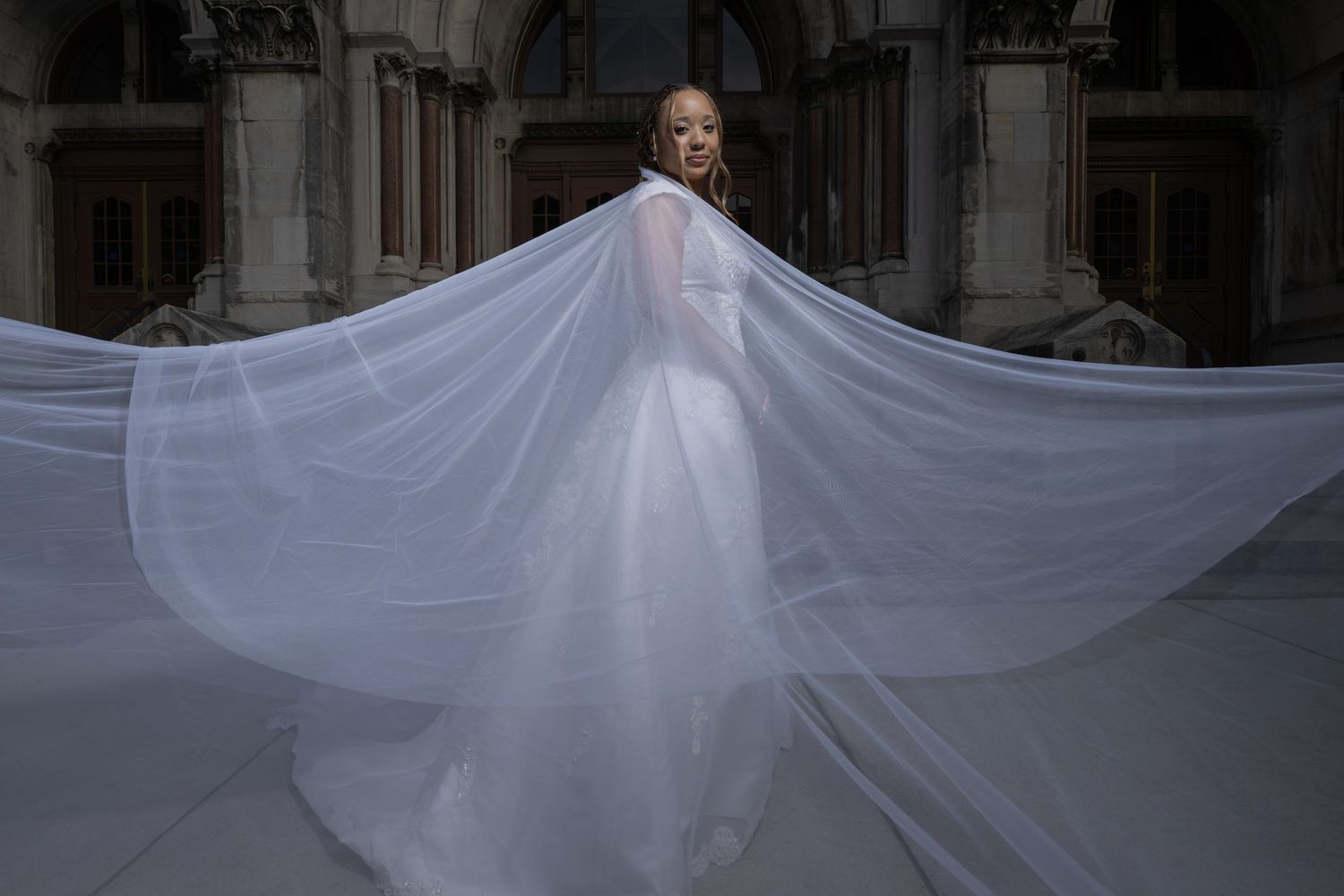 Bride in a flowing white wedding gown with a dramatic veil billowing in the wind, posed before grand stone columns.