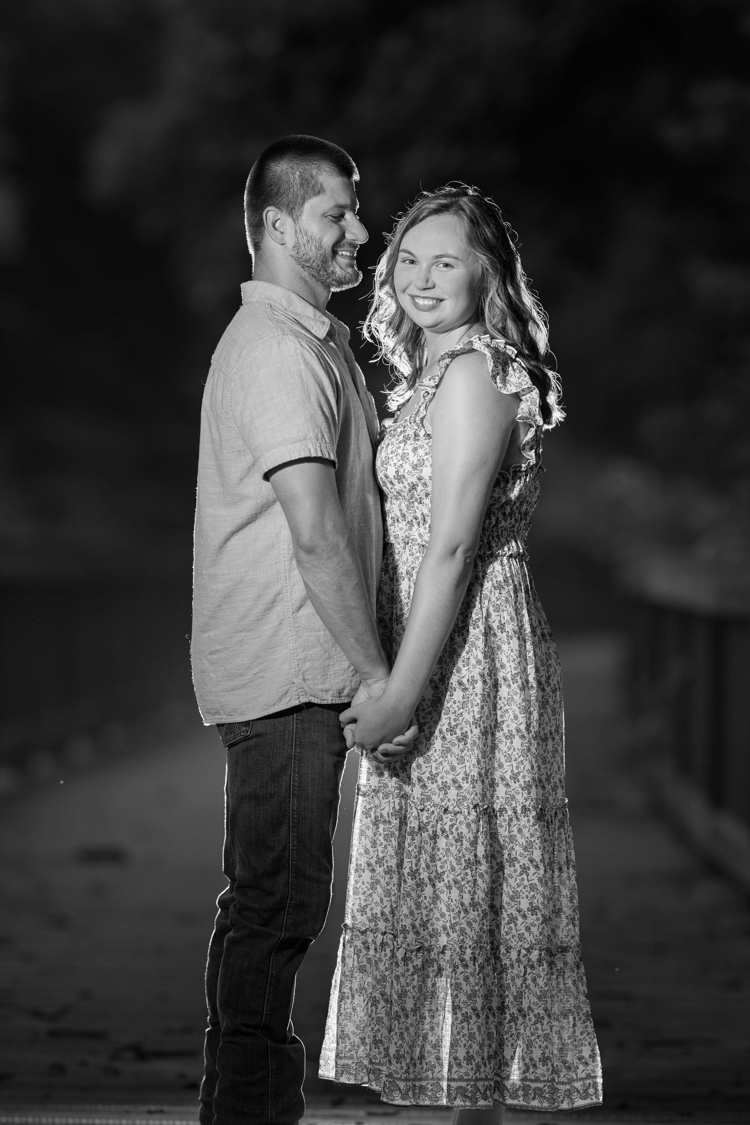 Black and white photo of a smiling couple holding hands outdoors at night, woman in floral dress, man in casual shirt.