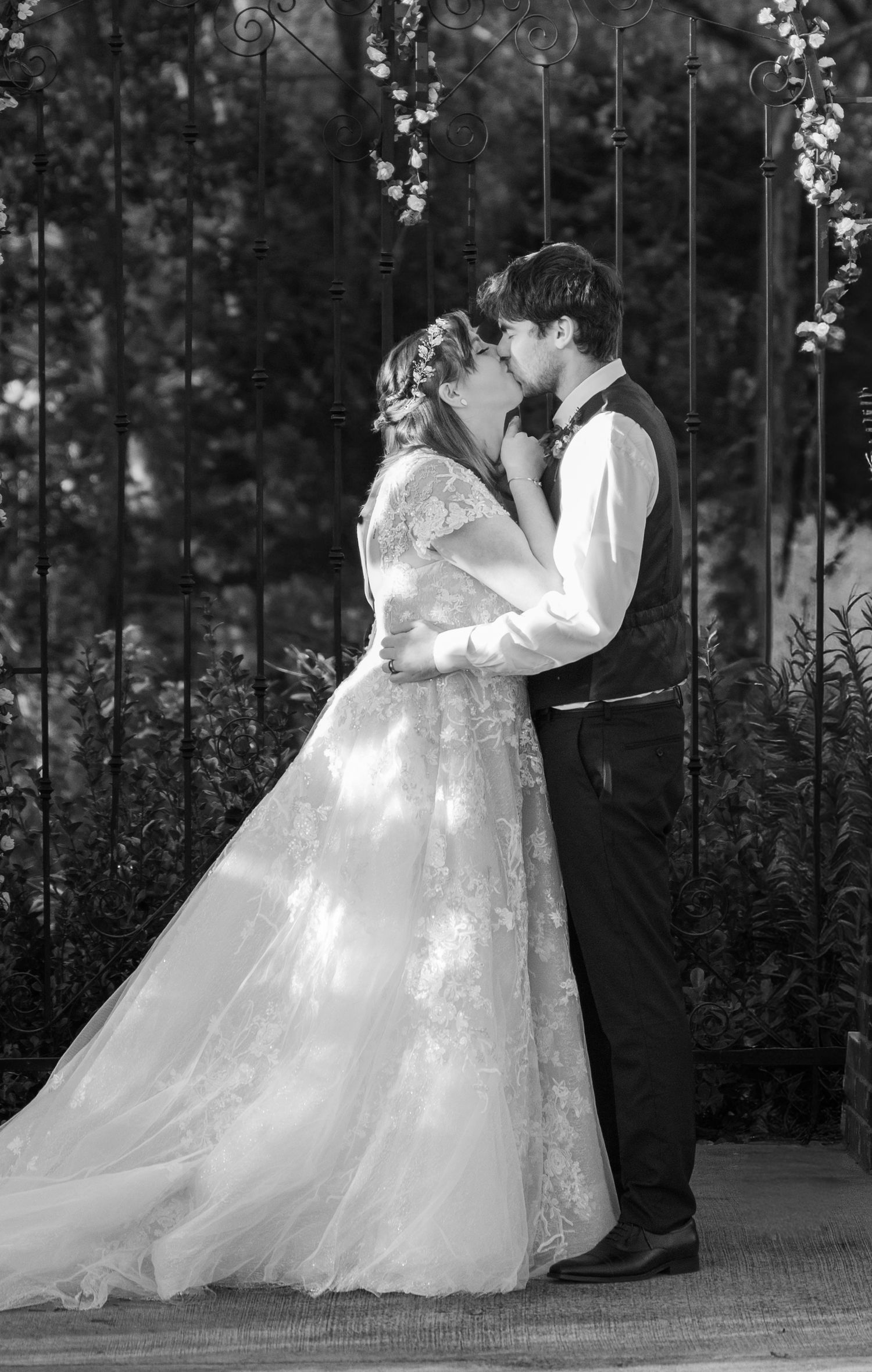 Black and white wedding photo of bride and groom sharing first kiss under floral arch in outdoor ceremony.