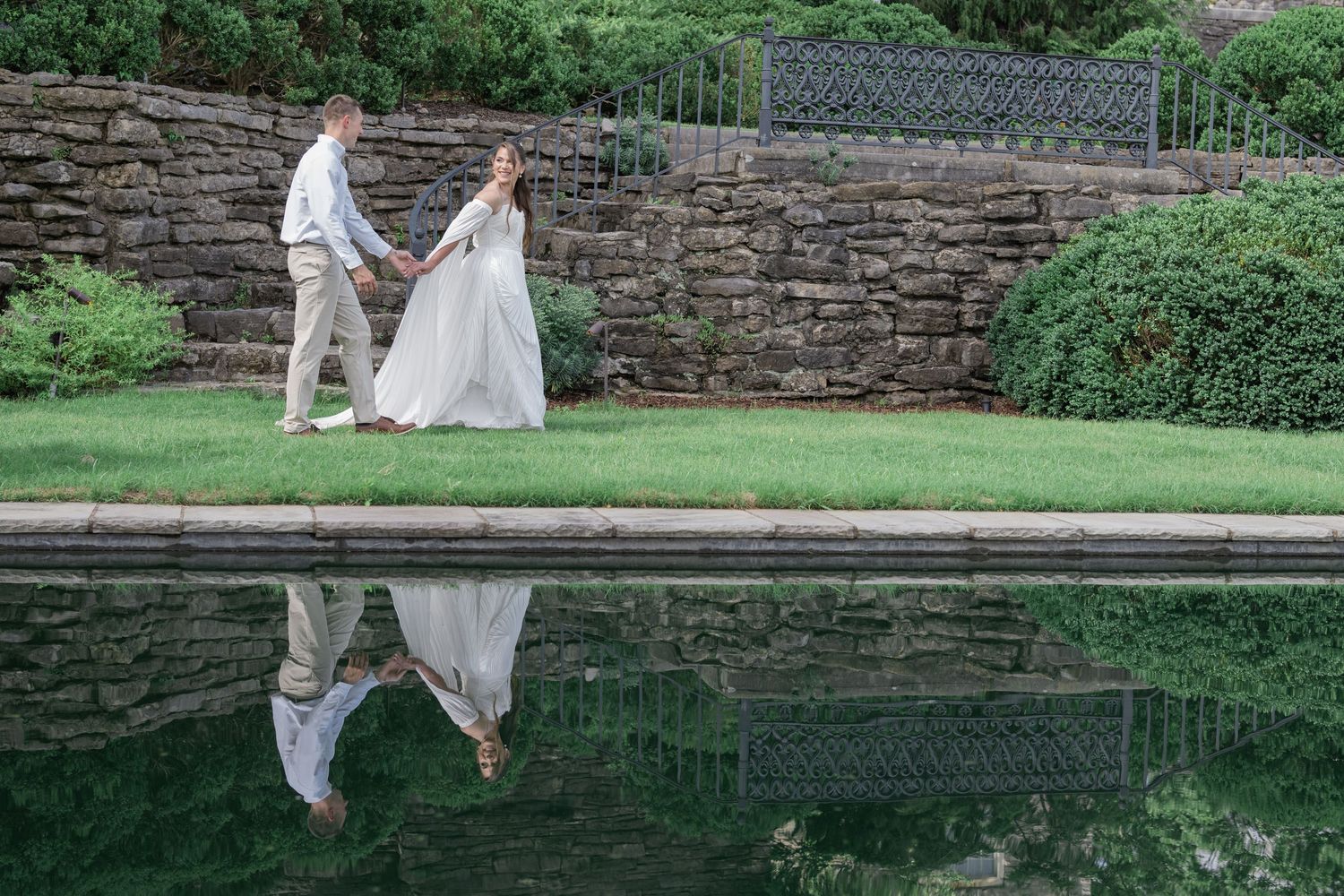 Bride and groom walking hand in hand beside a reflective pool with a stone wall backdrop at an outdoor wedding venue.