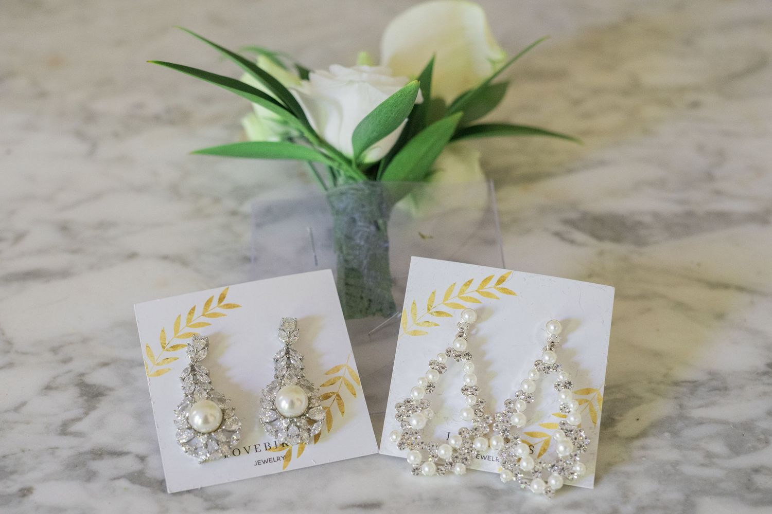 Crystal and pearl bridal drop earrings displayed on cards with white roses on marble.