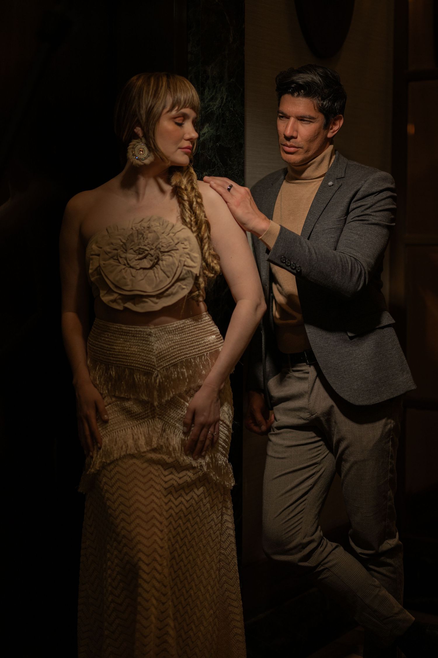 A man in a gray suit touches the shoulder of a woman in a gold fringe gown with a braided hairstyle.