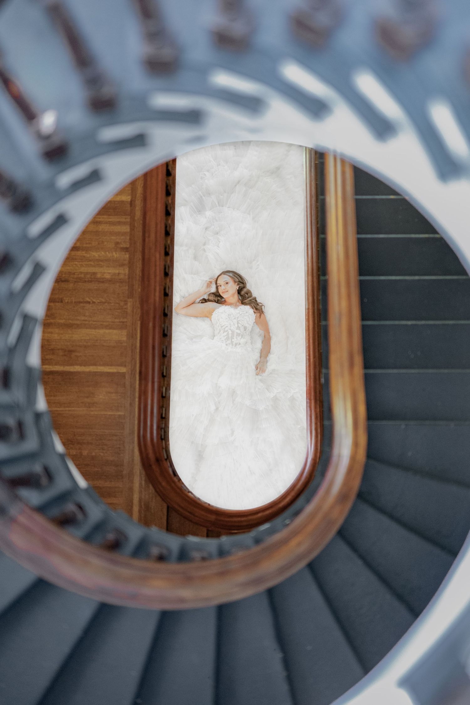 Aerial view of a bride in a white gown lying on marble floor, framed by an elegant spiral staircase.