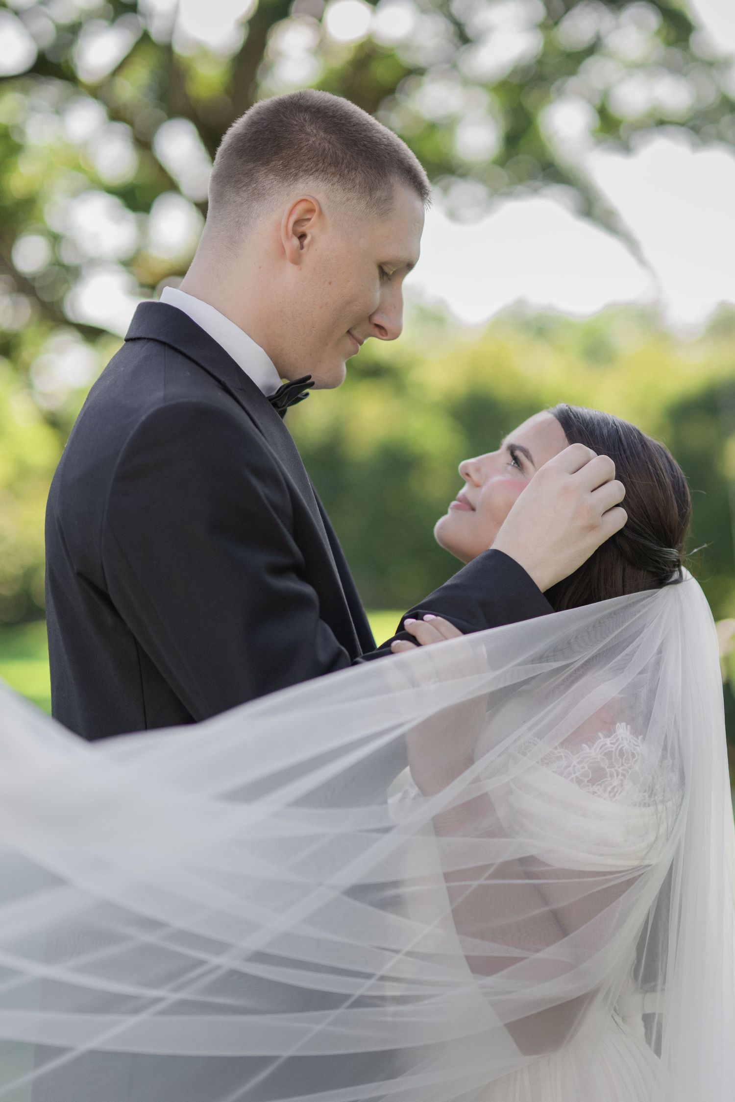 Bride and groom share a romantic moment outdoors, her veil flowing in the breeze as he gently holds her face.