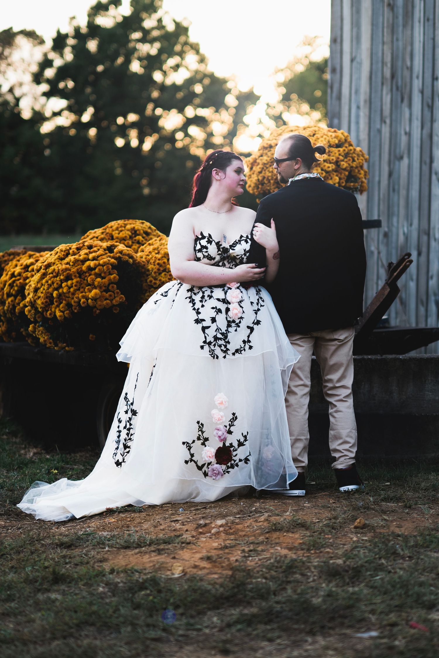 Bride in white gown with black floral lace and groom in black tuxedo pose near rustic barn with yellow mums at sunset.