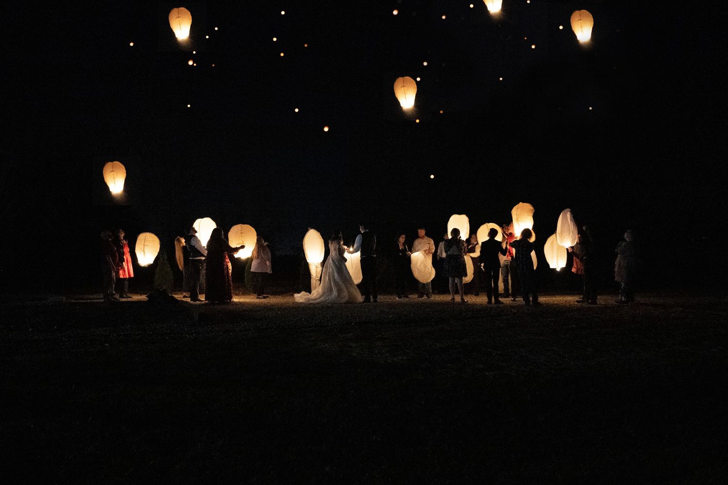 Wedding guests releasing glowing sky lanterns into the night sky during an outdoor evening celebration.
