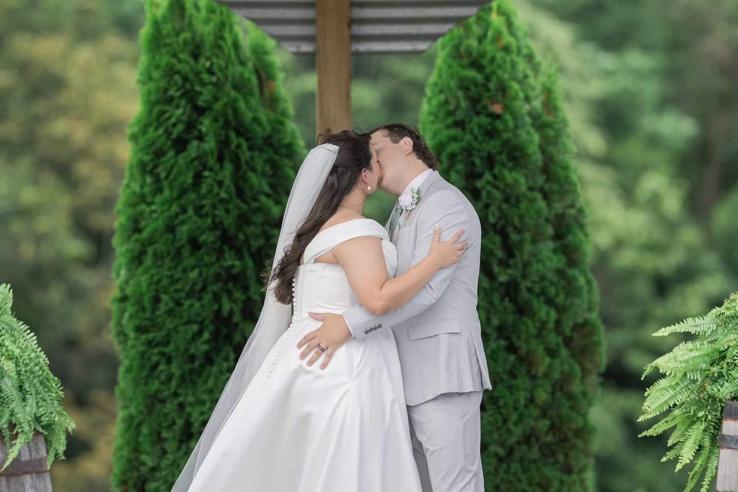 Bride and groom share their first kiss at outdoor wedding ceremony surrounded by tall evergreen trees.