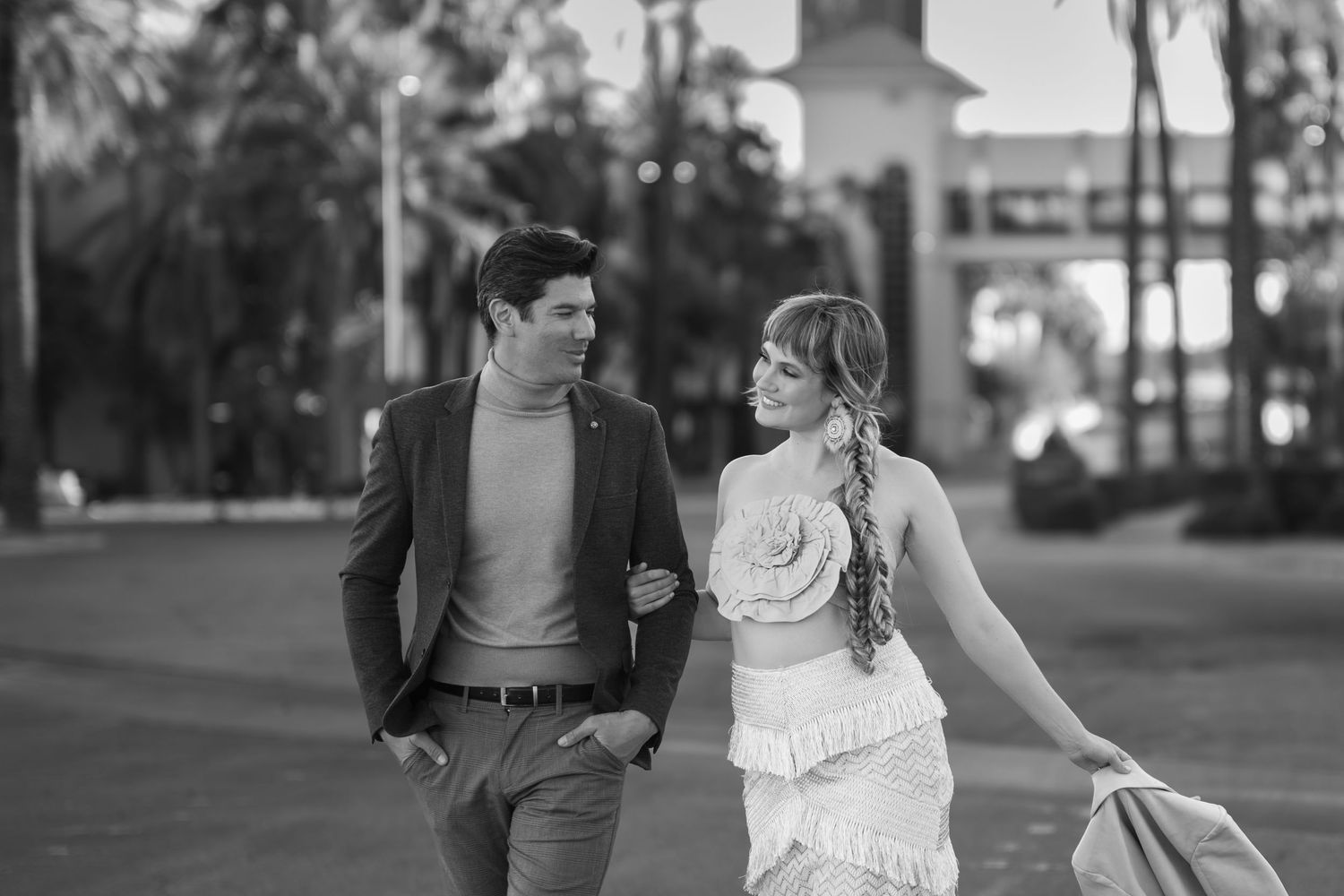 Black and white photo of an elegant couple walking together outdoors, woman in floral dress, man in blazer.