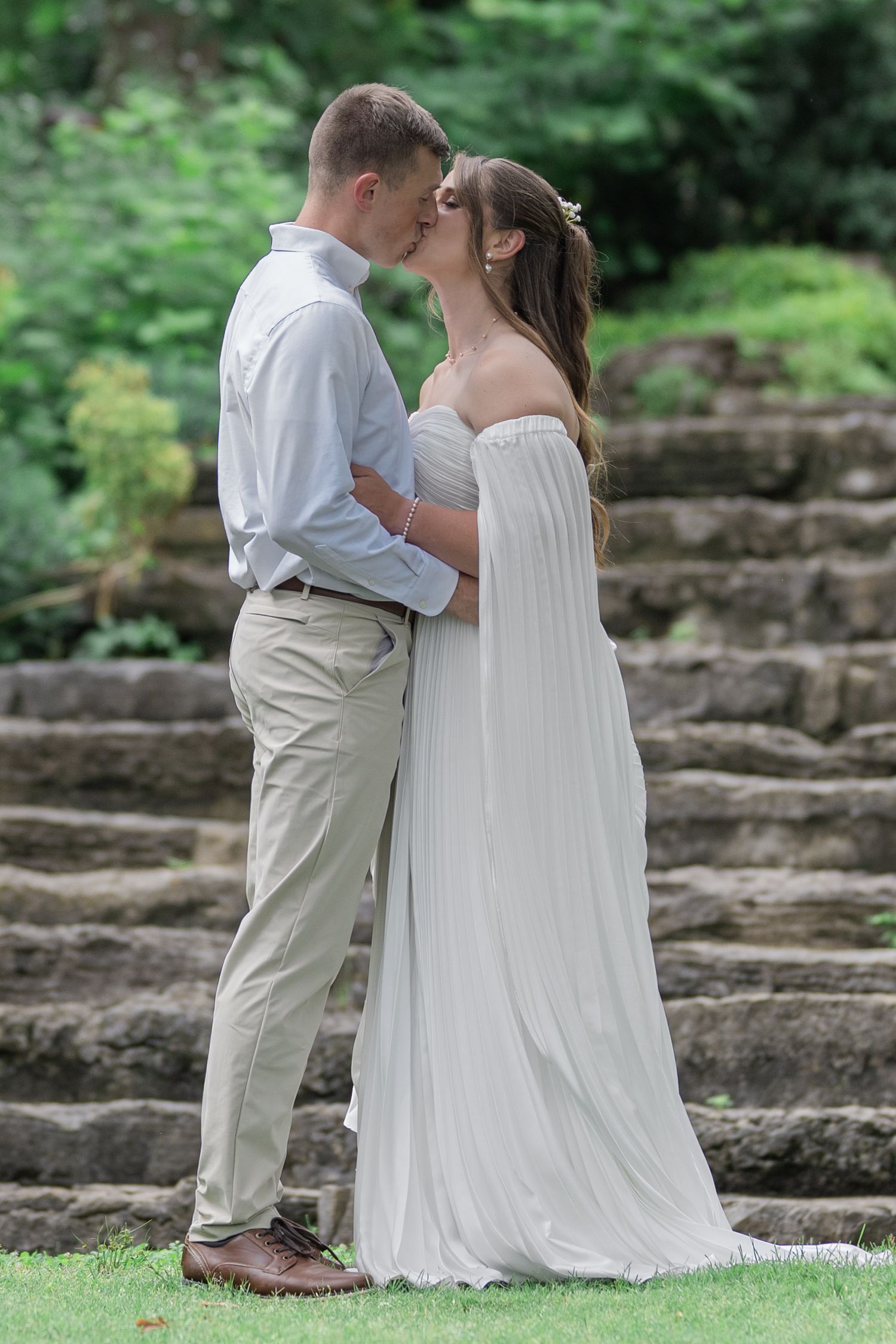 Bride and groom sharing a romantic kiss outdoors on stone steps surrounded by lush green garden foliage.
