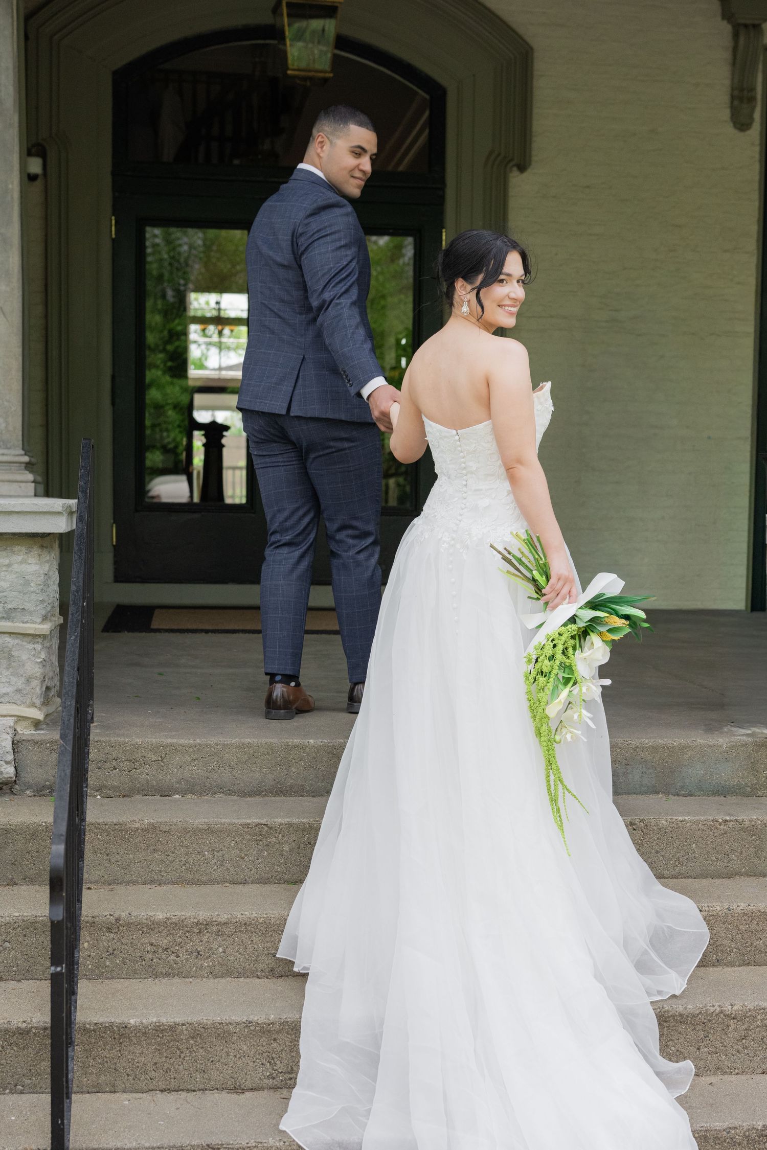 Bride in white strapless gown holding bouquet smiles while groom in navy suit leads her up stone steps.