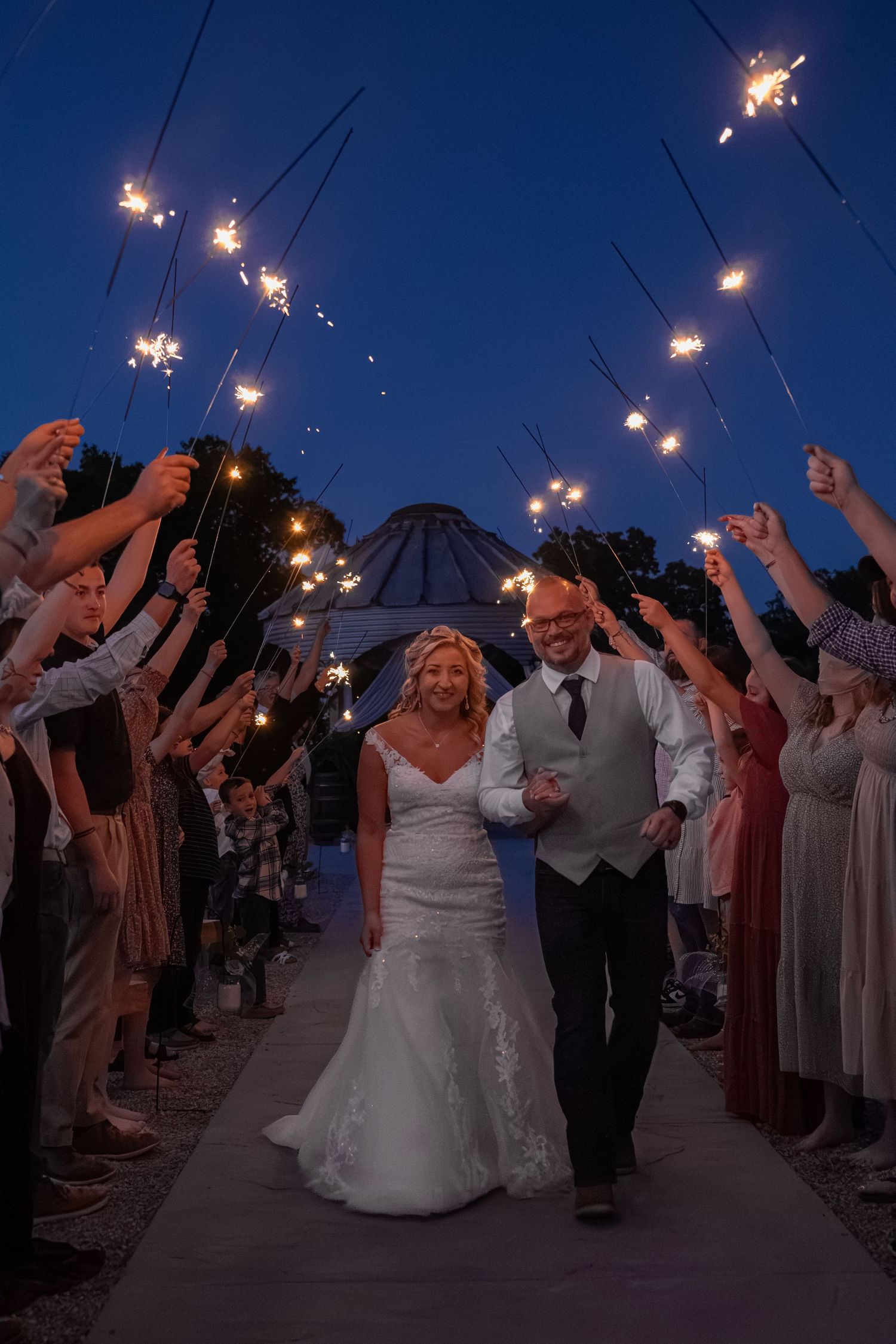 Bride and groom walk through sparkler tunnel at dusk during outdoor wedding reception exit.