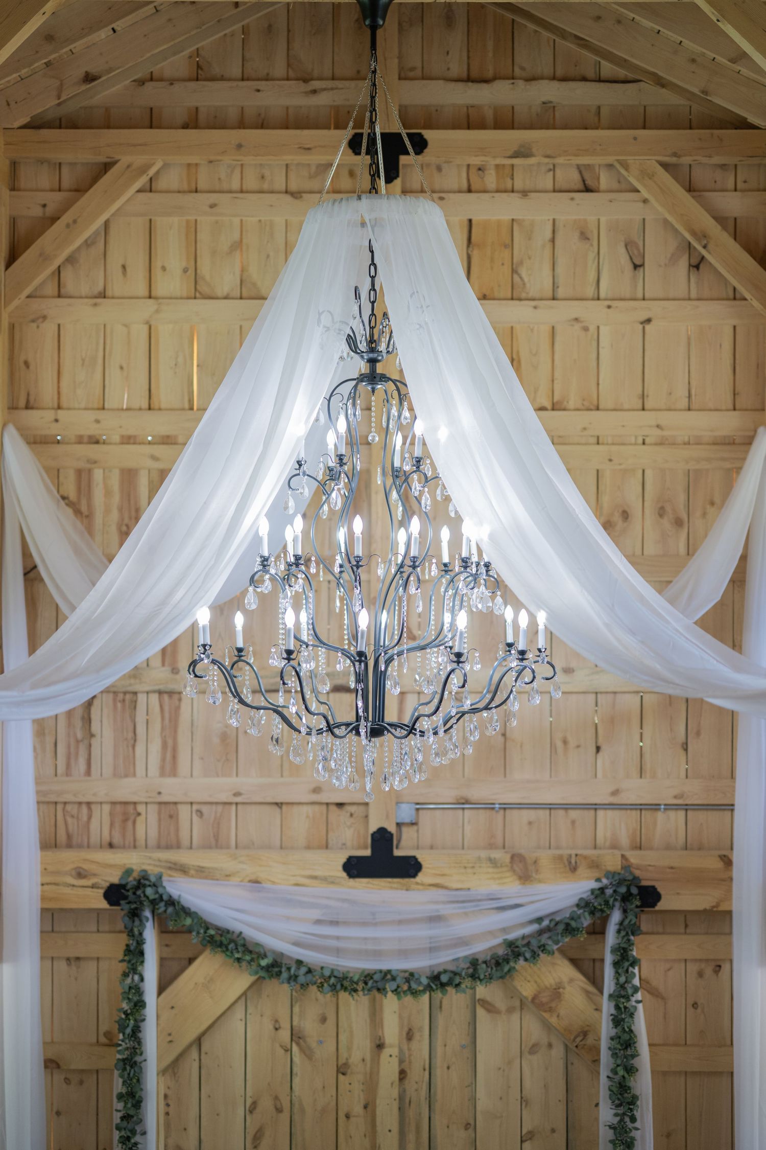 Elegant crystal chandelier with candle lights draped in white fabric hanging in a rustic wooden barn wedding venue.