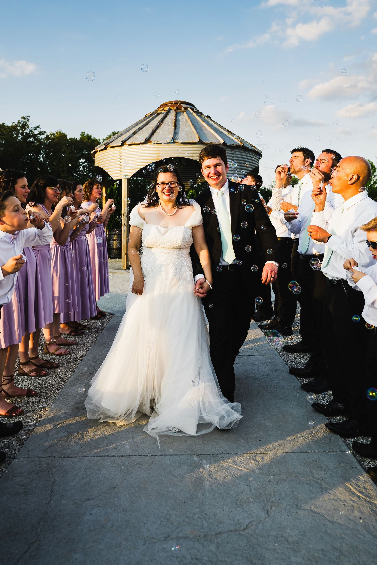 A smiling bride and groom walk through a bubble-filled exit lined with guests and lavender-dressed bridesmaids.