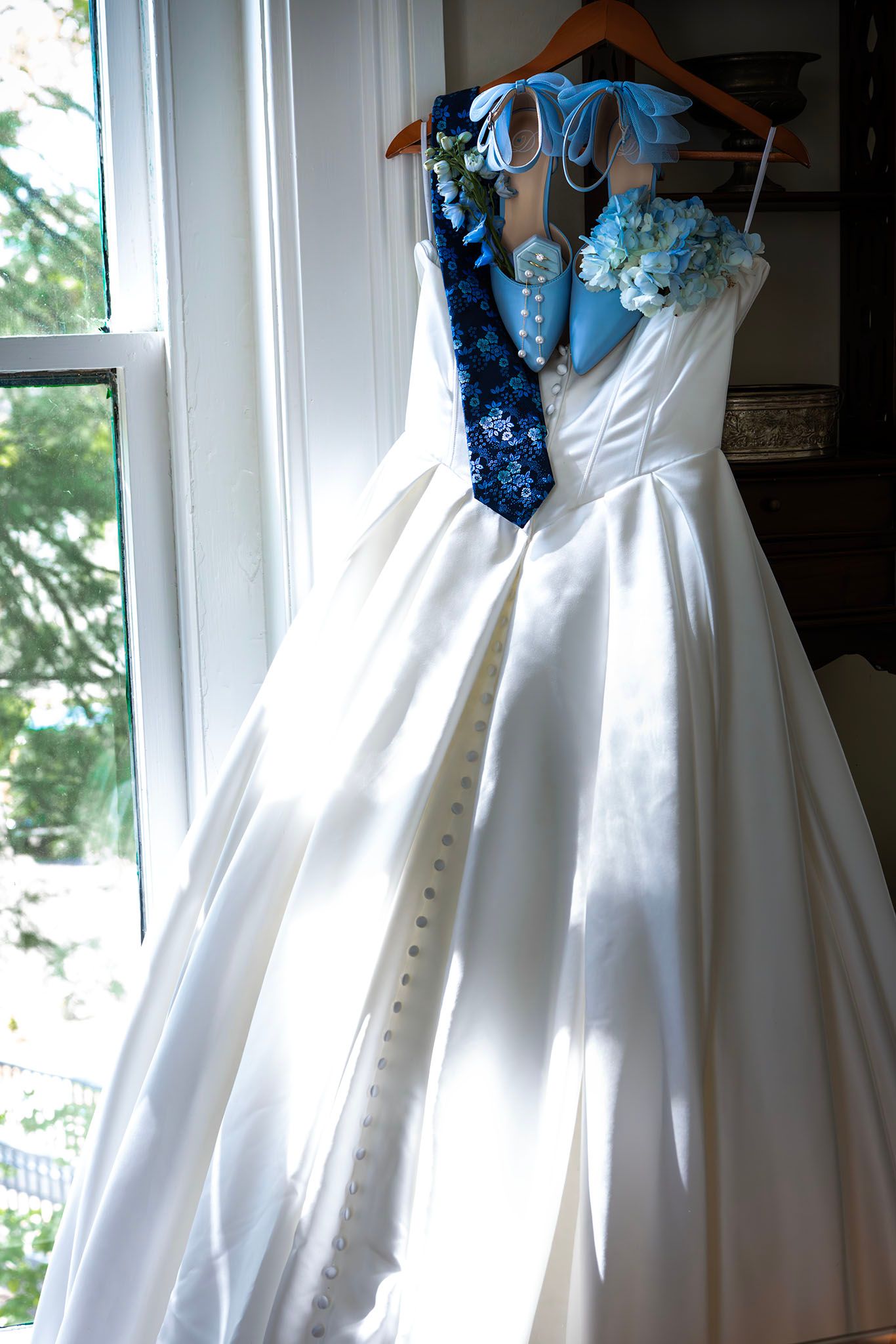 White satin ballgown wedding dress hanging by window with blue floral accessories and navy tie on wooden hanger.