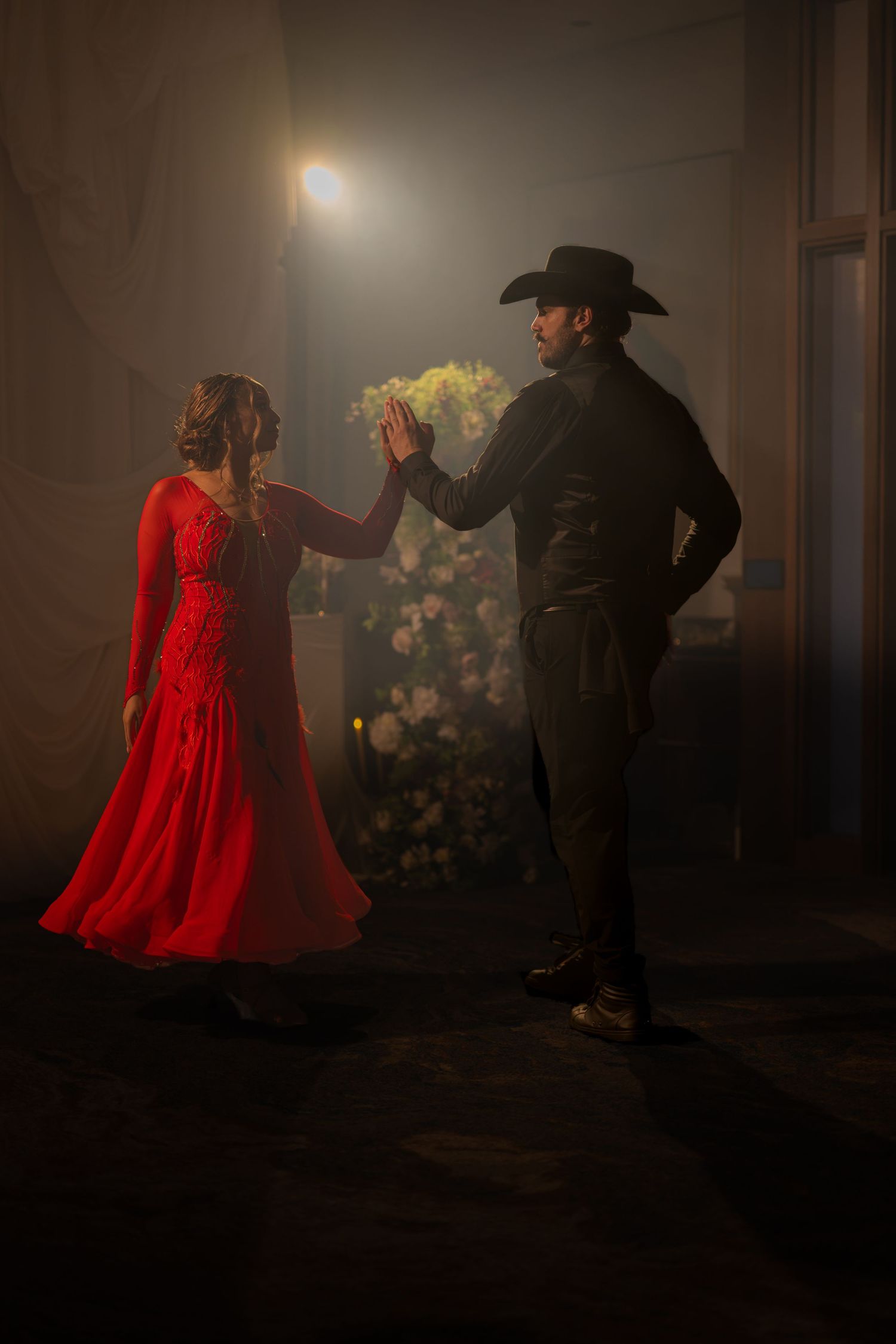 A couple dances in dim light, woman in red gown, man in black cowboy hat, with flowers glowing behind them.