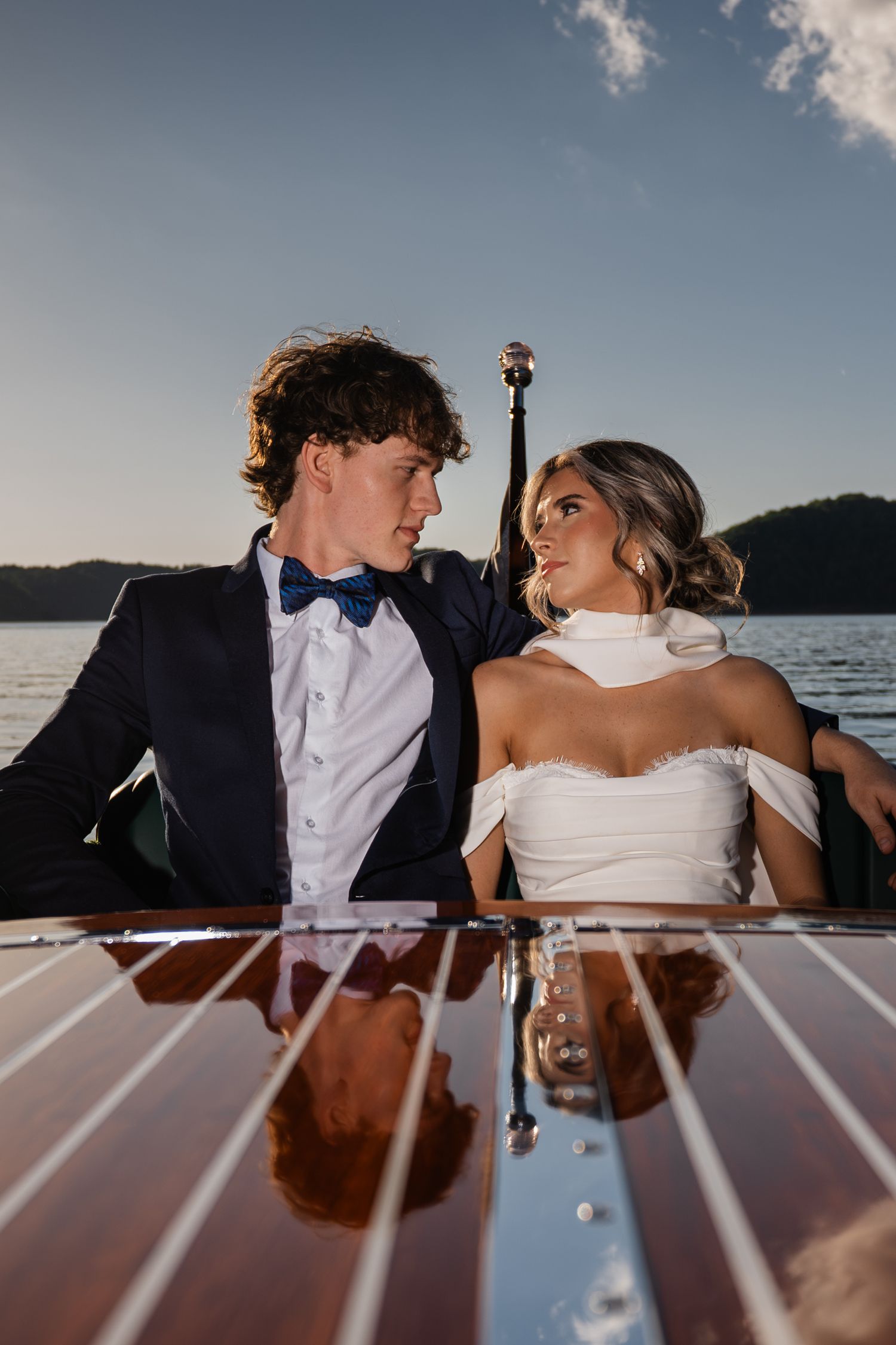 Elegant couple in wedding attire sharing a romantic moment on a classic wooden boat at sunset by the water.
