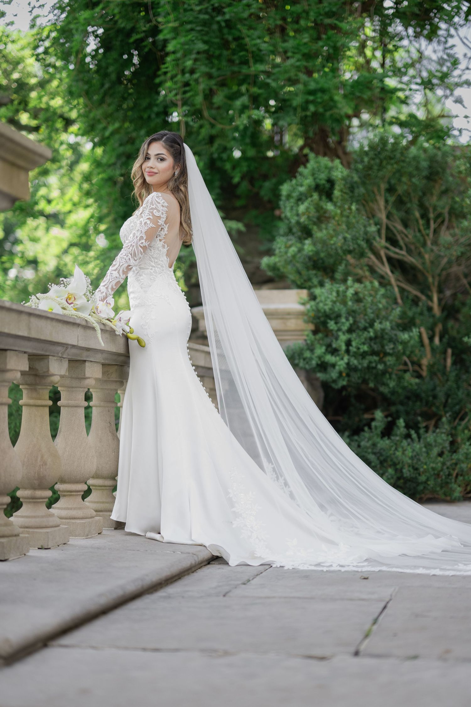 Elegant bride in a fitted white lace gown with long veil poses by stone balustrade in lush garden setting.