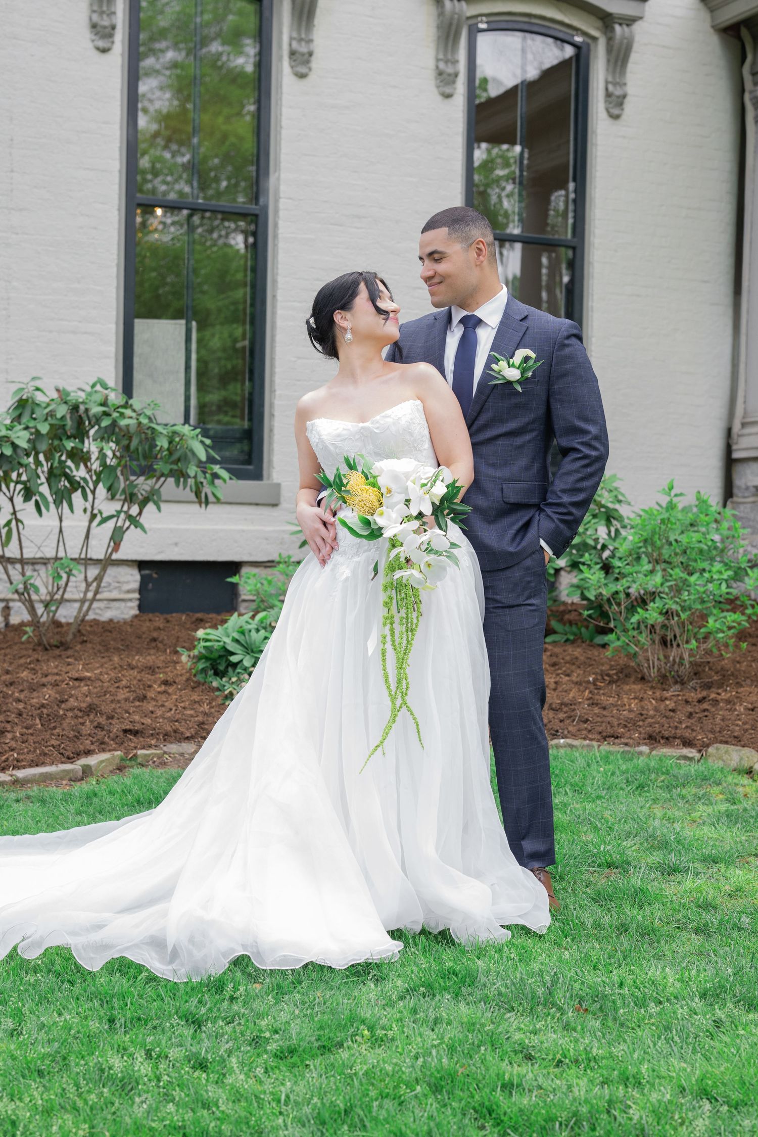Bride in white strapless gown with bouquet and groom in navy suit share a romantic moment on green lawn outside elegant venue.