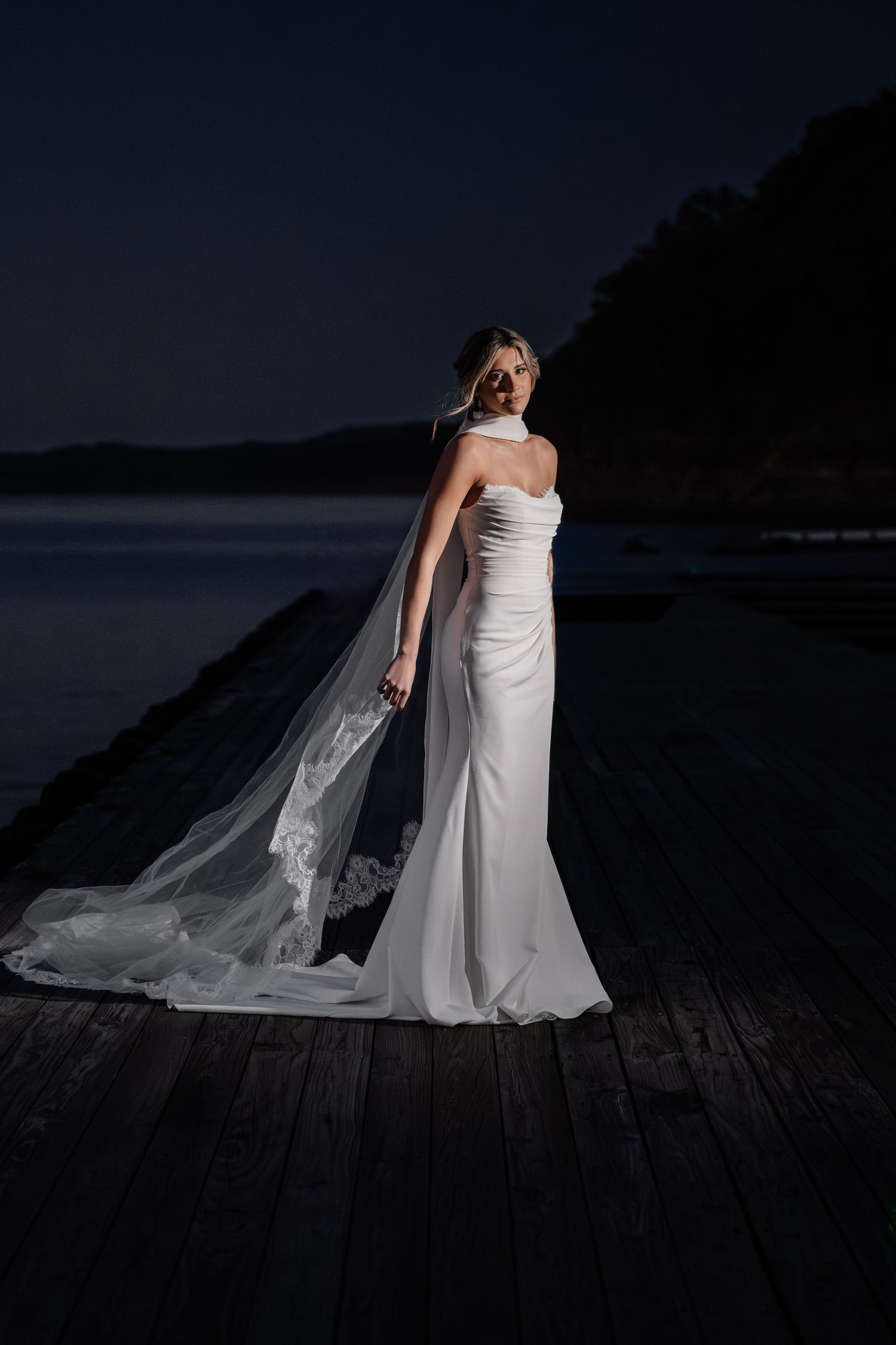 Bride in white fitted gown with lace veil poses on dark waterfront dock at night.