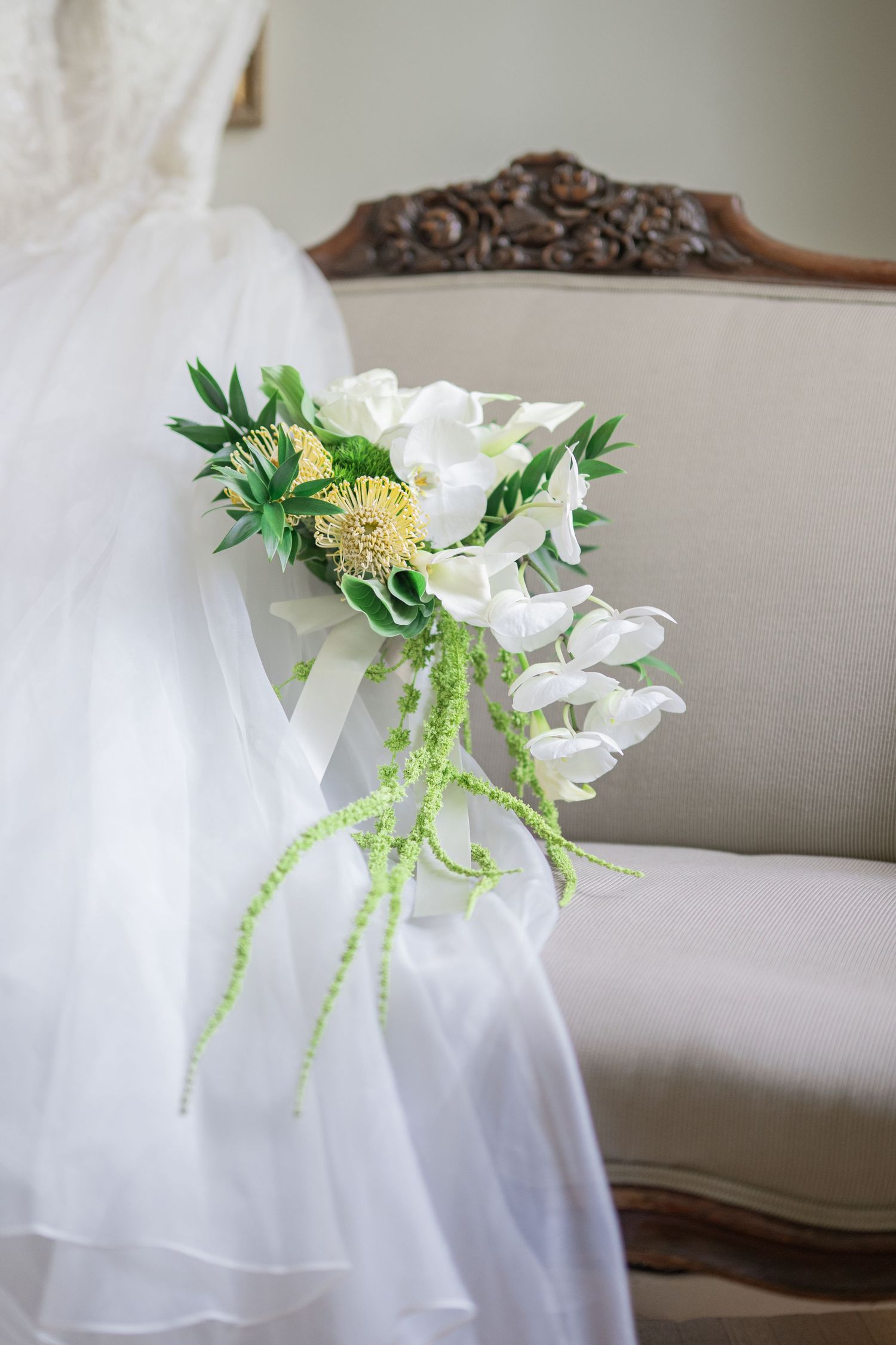 Bride holding white orchid and green cascading wedding bouquet in front of elegant vintage sofa.