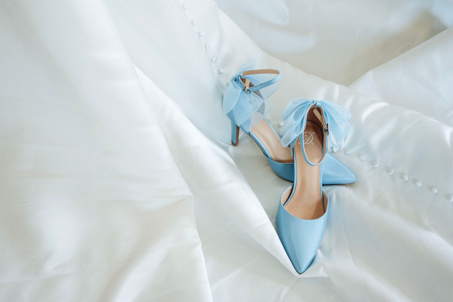 Light blue bridal heels with bow detail resting on white wedding dress fabric.