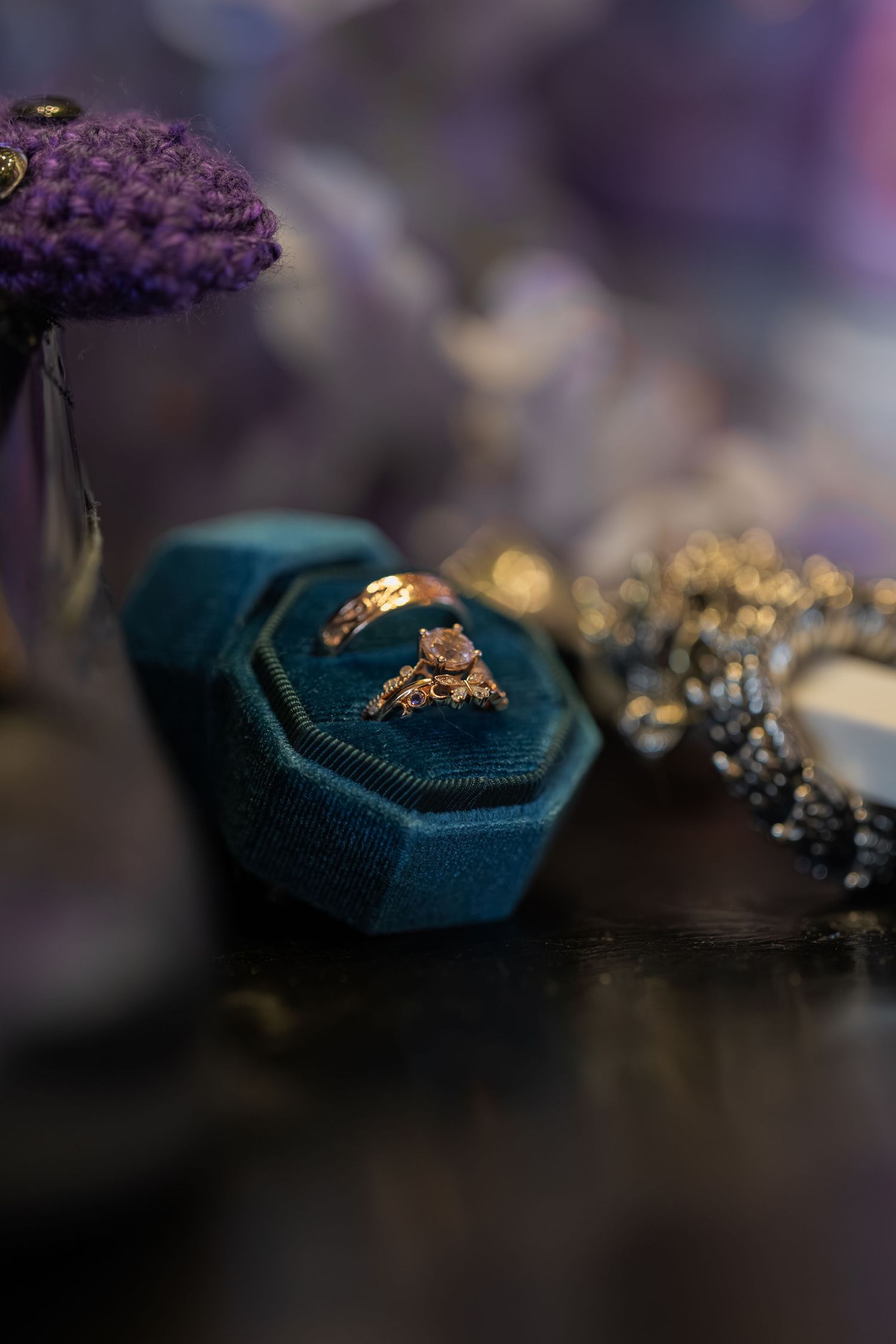 Gold engagement ring with pink stone in teal velvet ring box, surrounded by purple flowers and jewelry.