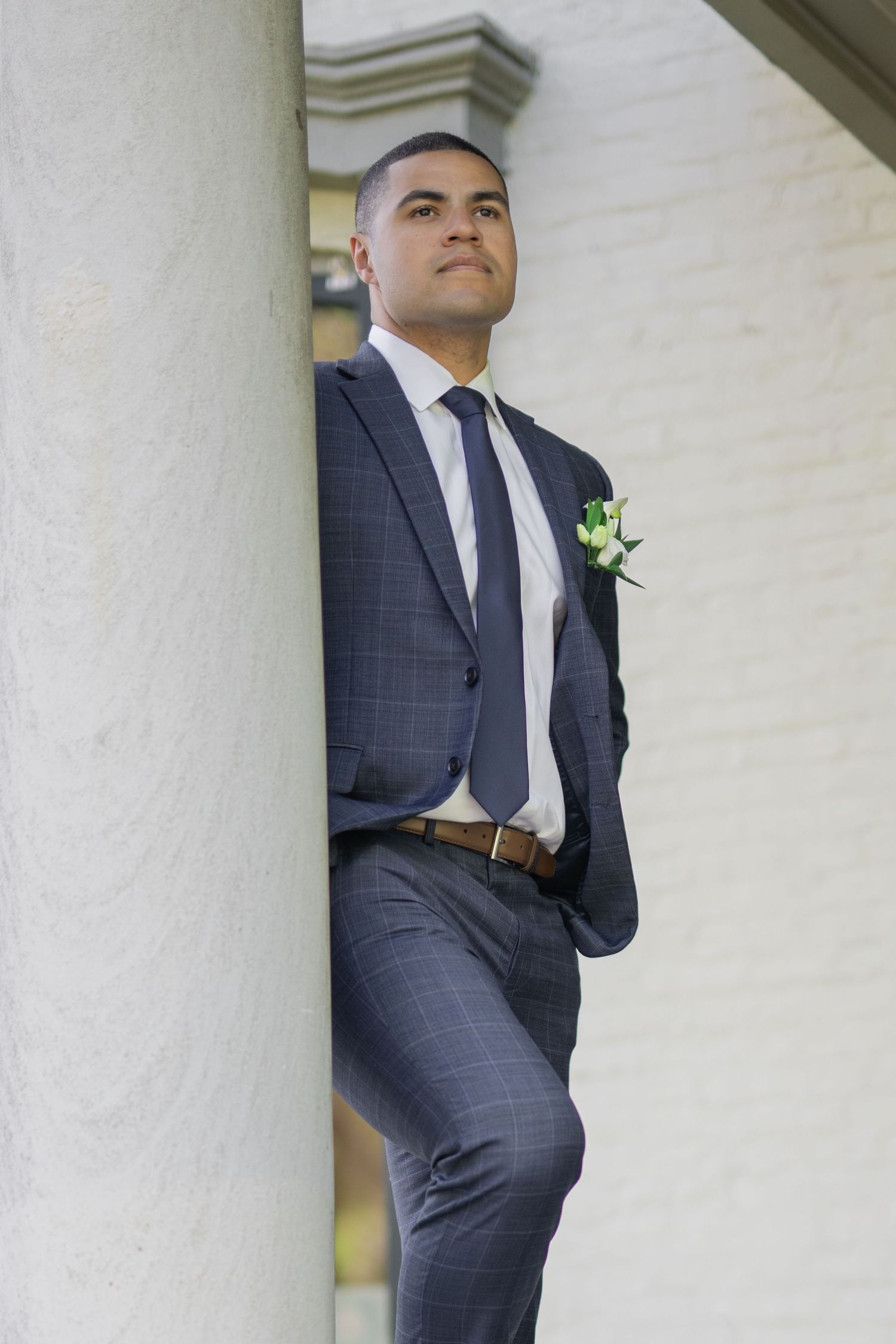 A well-dressed groom in a navy plaid suit with boutonniere leans against a white column outdoors.